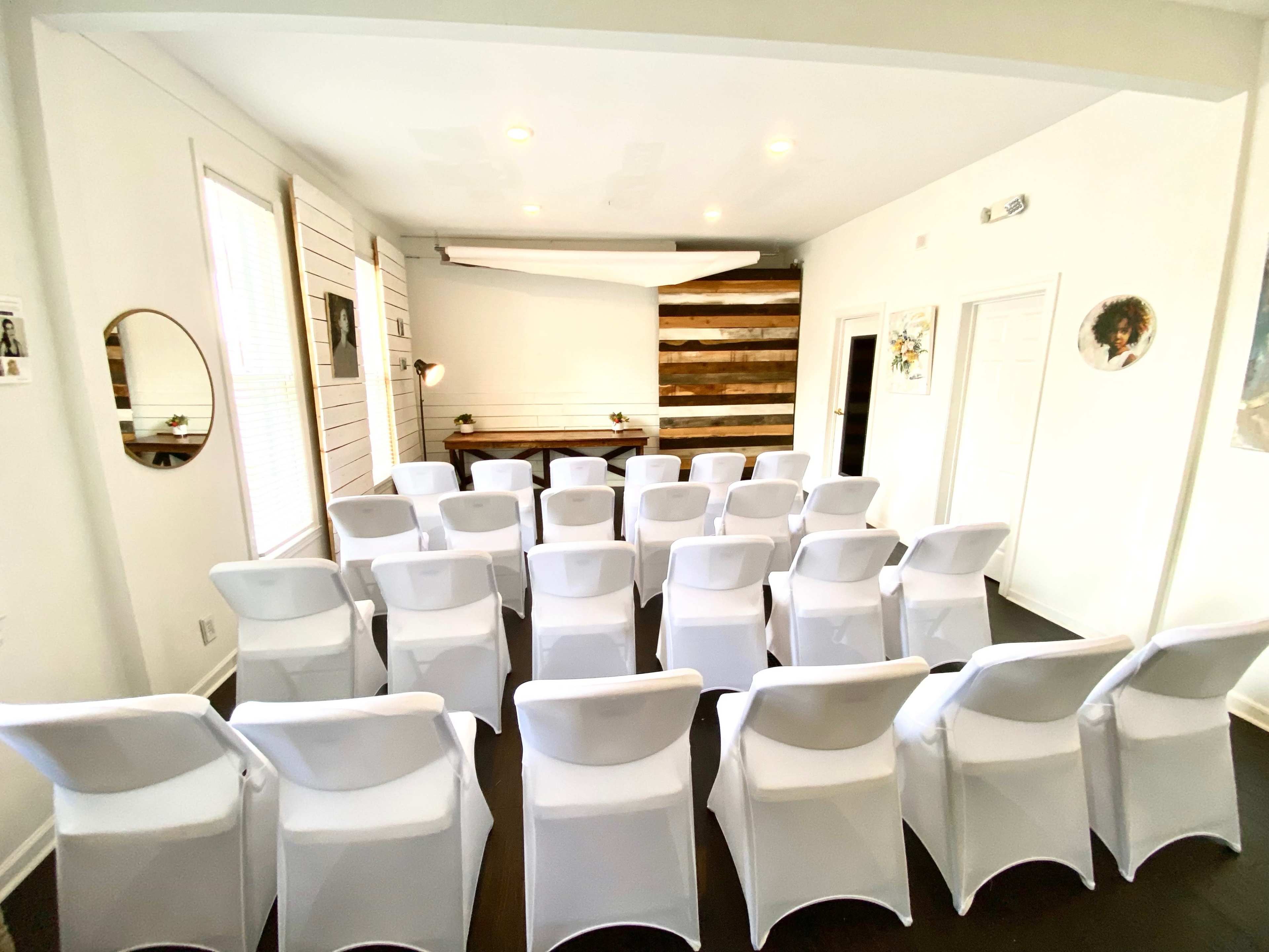 A row of white chairs is set up facing a wooden-paneled wall in a well-lit room.