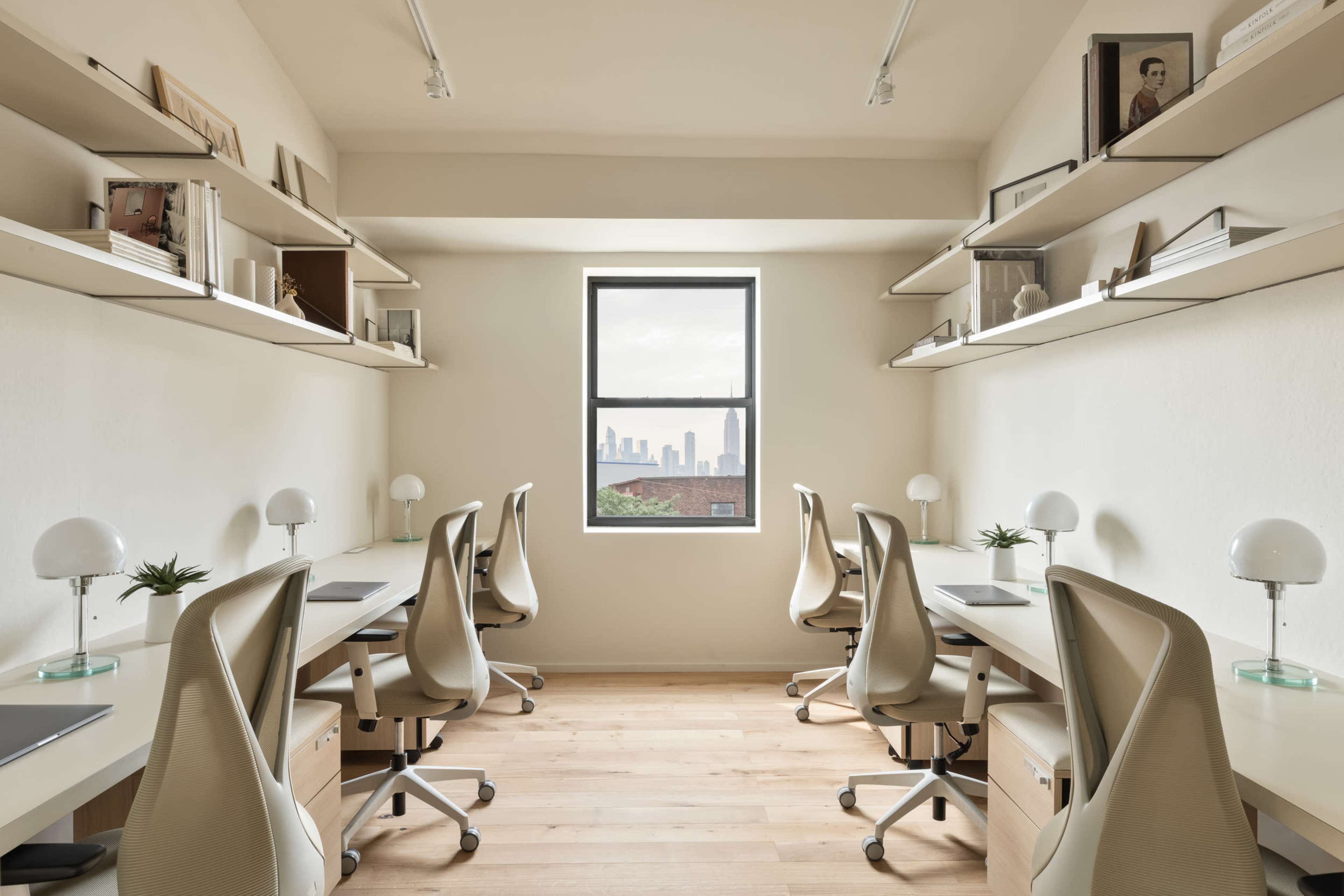 The image shows a modern office space with four workstations, each equipped with a chair and desk lamp, and large shelves holding books and decorative items, all set against a window revealing a city skyline.