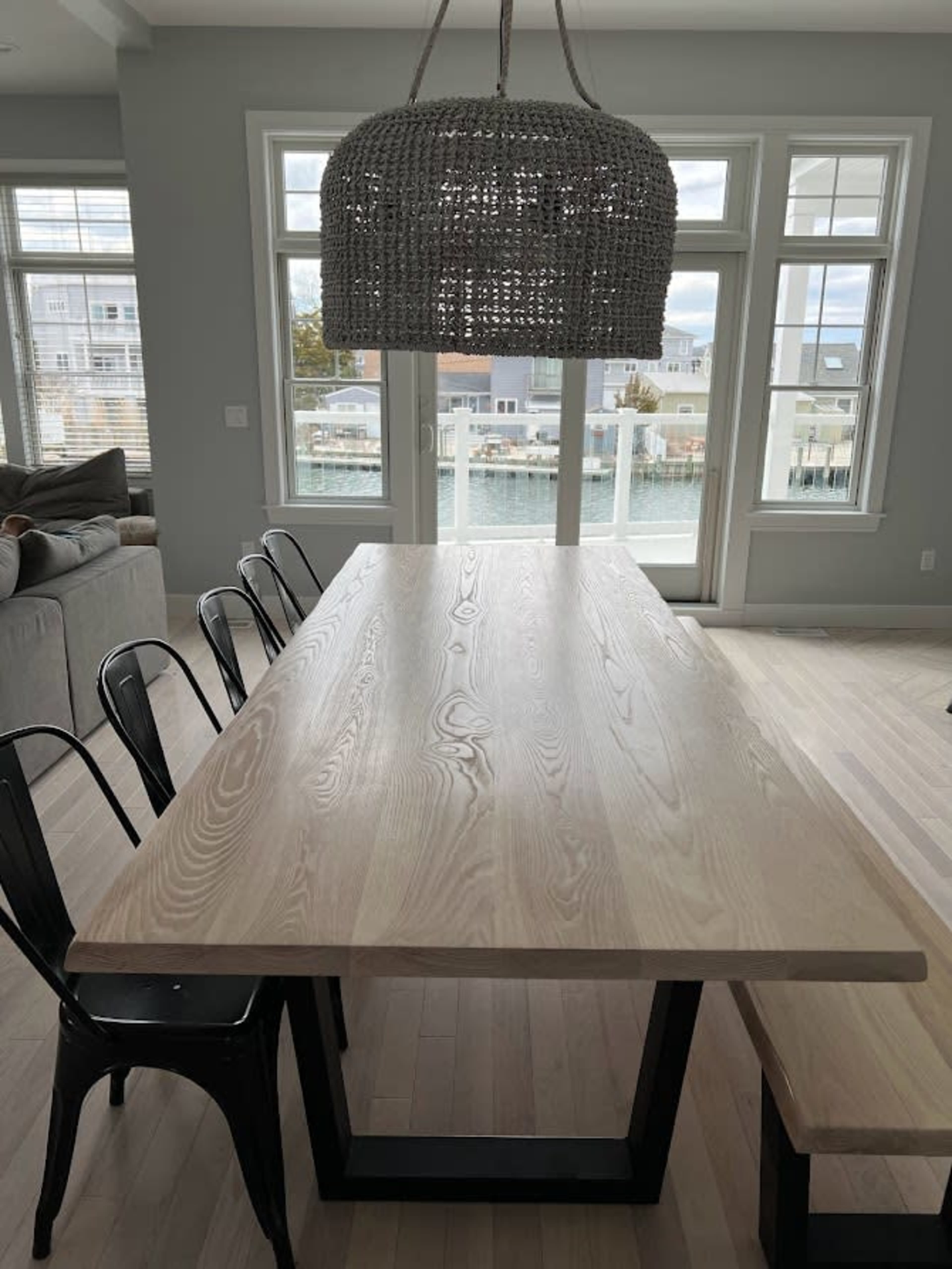 A large wooden dining table with black metal chairs is positioned near a window overlooking a waterfront view.