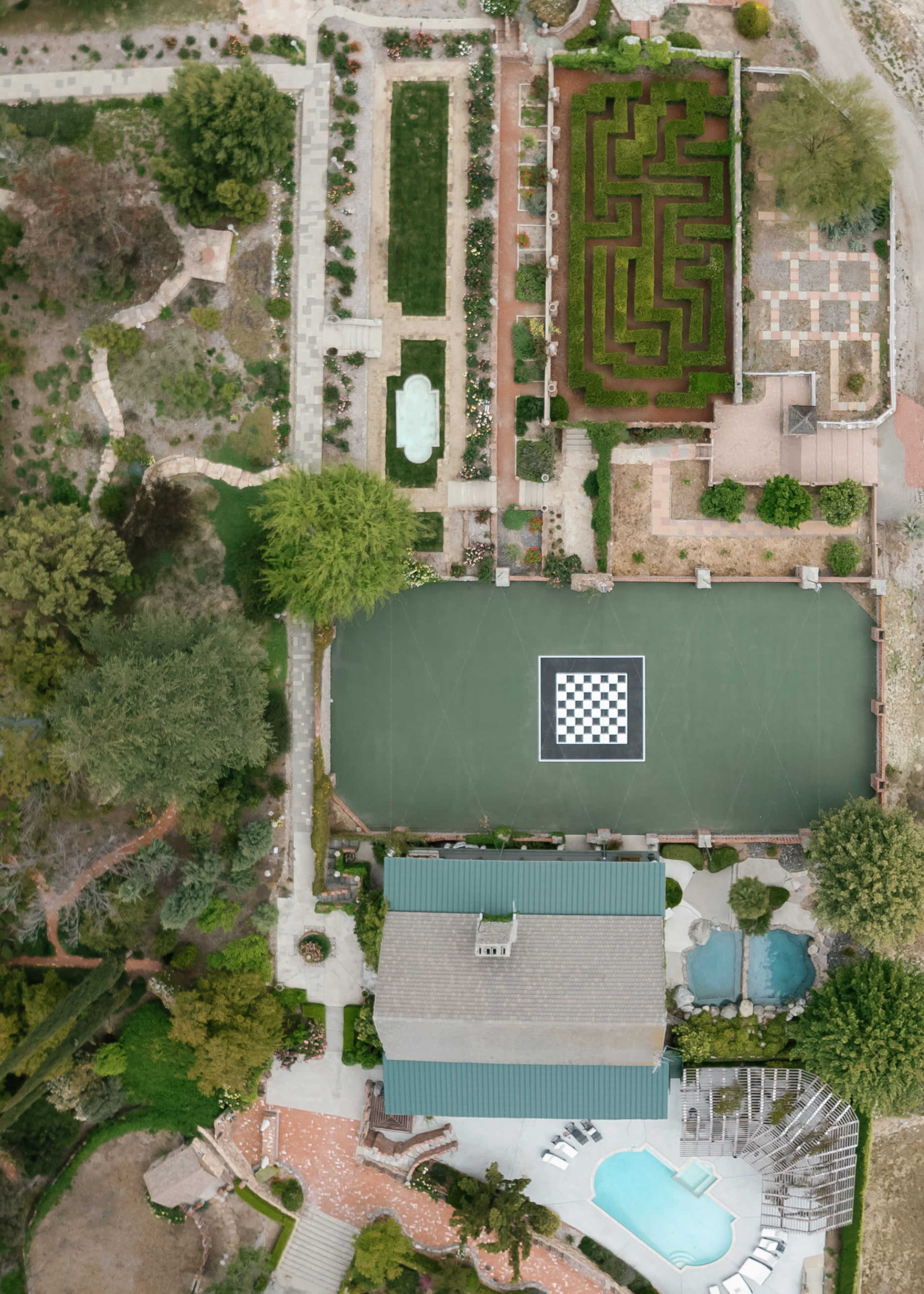The image shows an aerial view of a landscaped property featuring a maze garden, a swimming pool, and a large chessboard layout on a court.