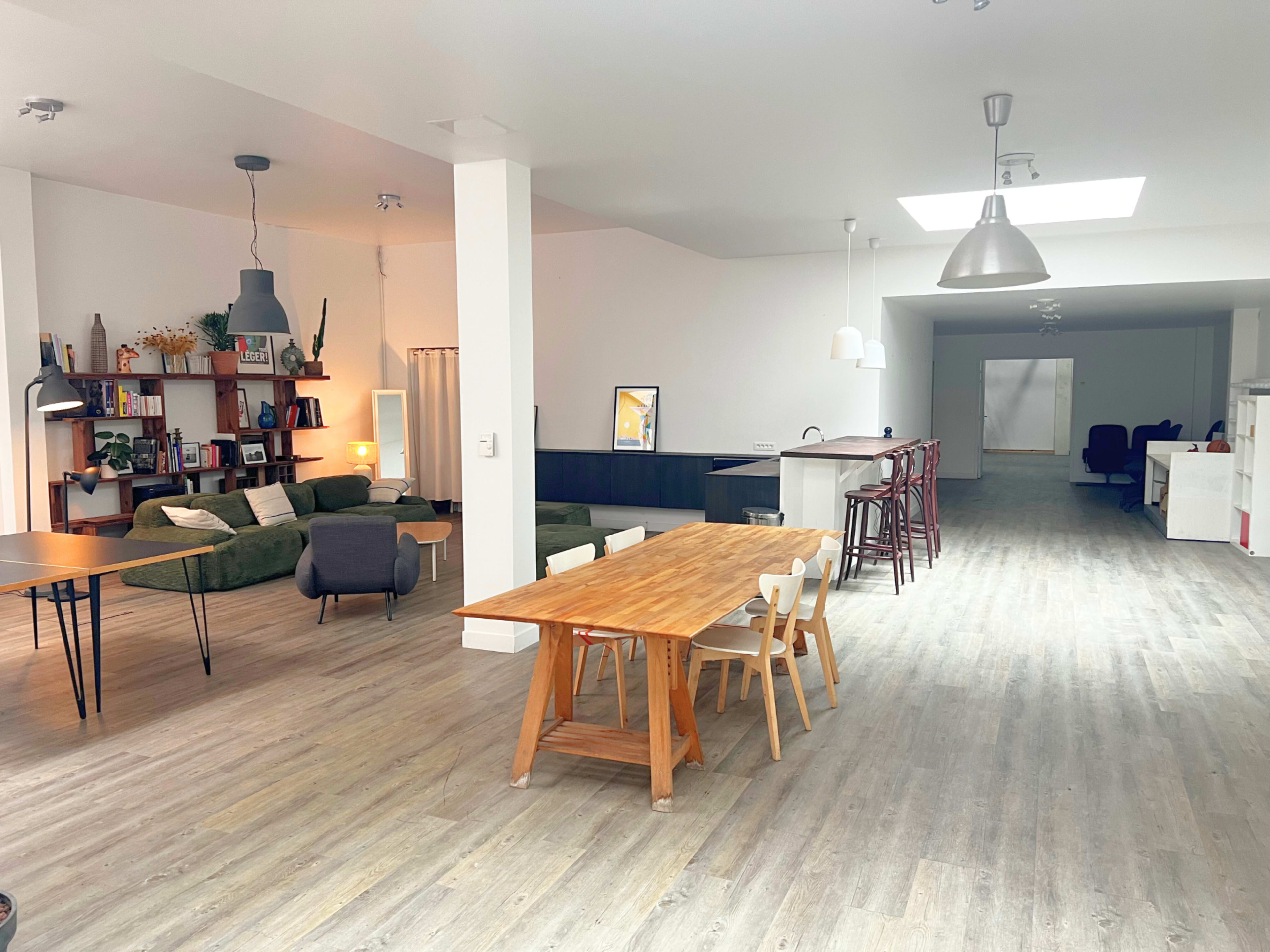 The image shows a spacious, minimalistic room with wooden flooring, featuring a dining table, seating area, and a kitchen in an open layout.