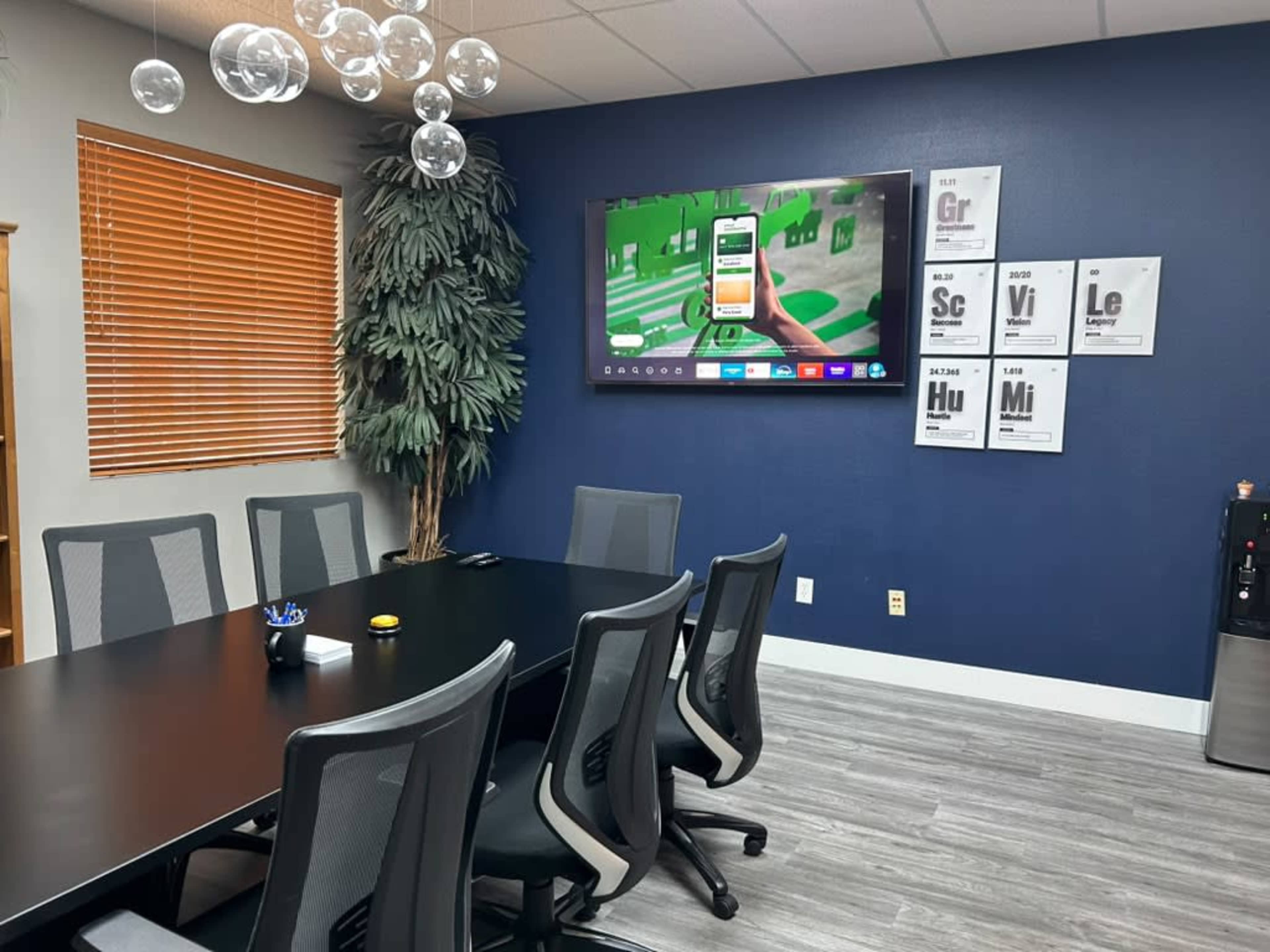 The image shows a modern conference room with a large table, several ergonomic chairs, a wall-mounted television displaying a colorful graphic, a potted plant in the corner, and framed posters on the wall.
