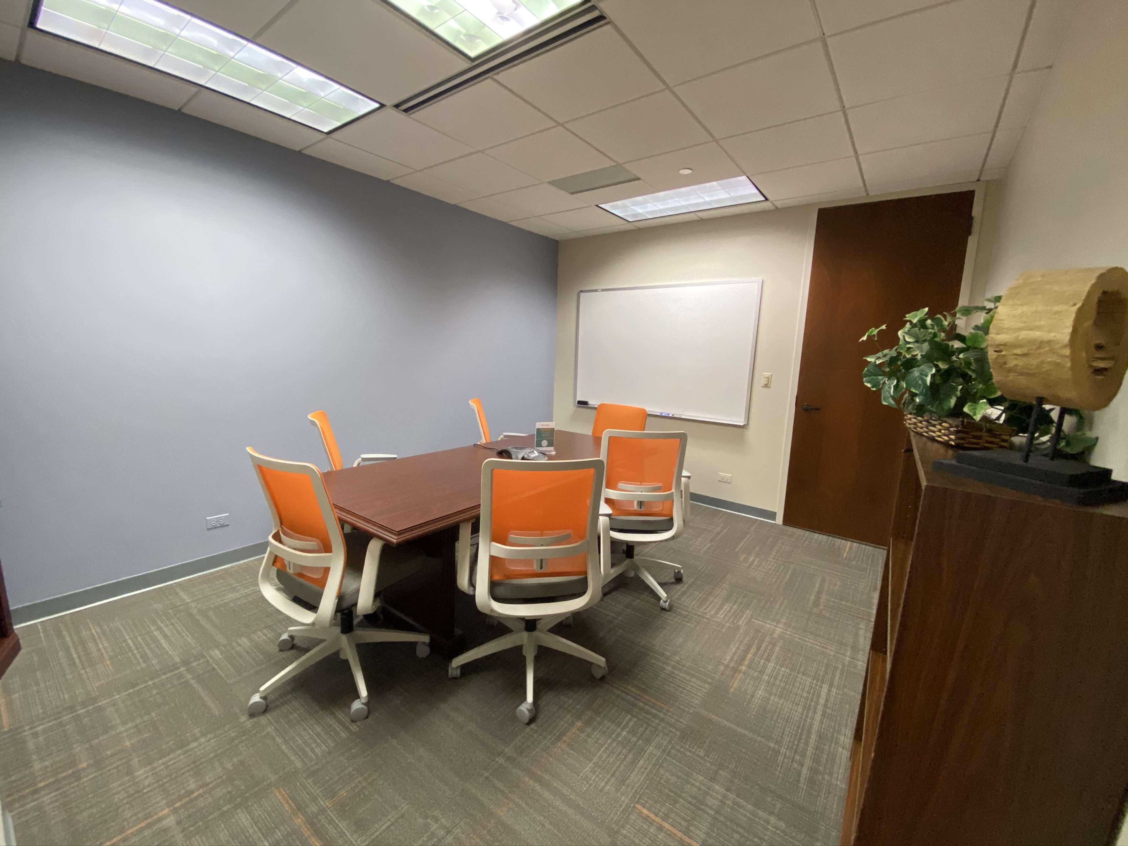A conference room features a rectangular table surrounded by orange and white chairs, with a whiteboard on one wall and a plant in a pot.