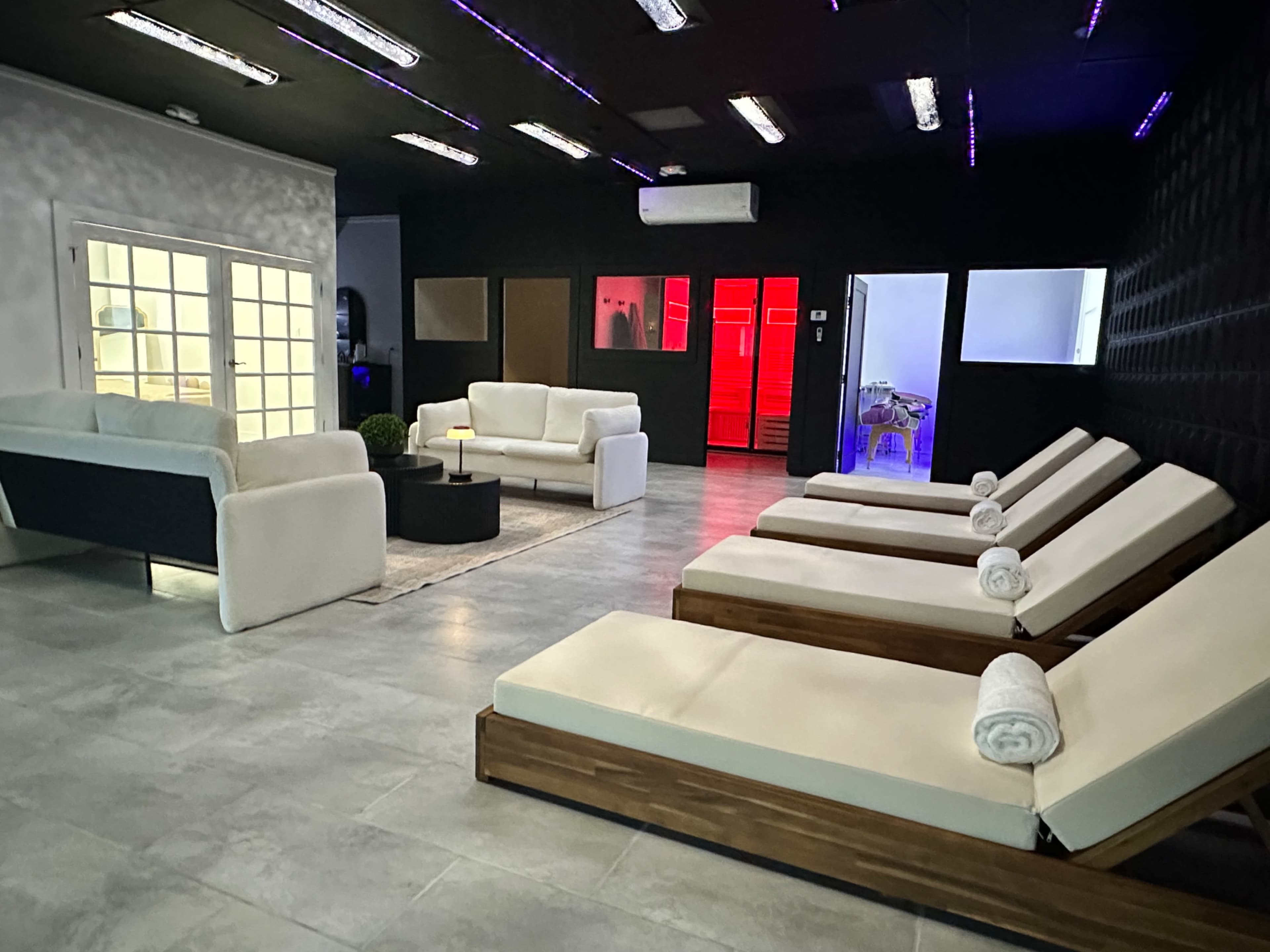 A modern relaxation area featuring white sofas, lounge chairs with rolled towels, and ambient lighting.