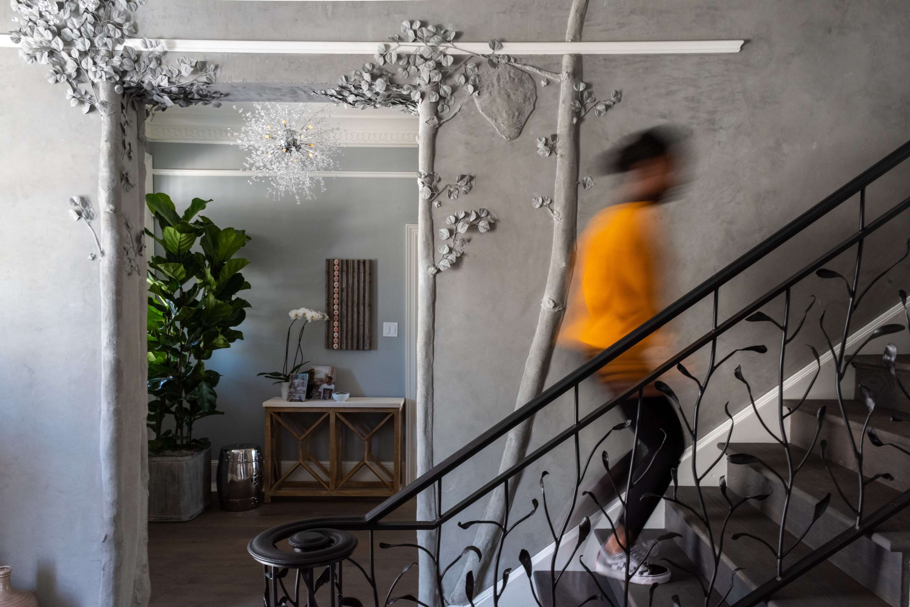 A person in a yellow jacket walks up a staircase in a modern entryway decorated with abstract tree-like wall sculptures and a large plant.