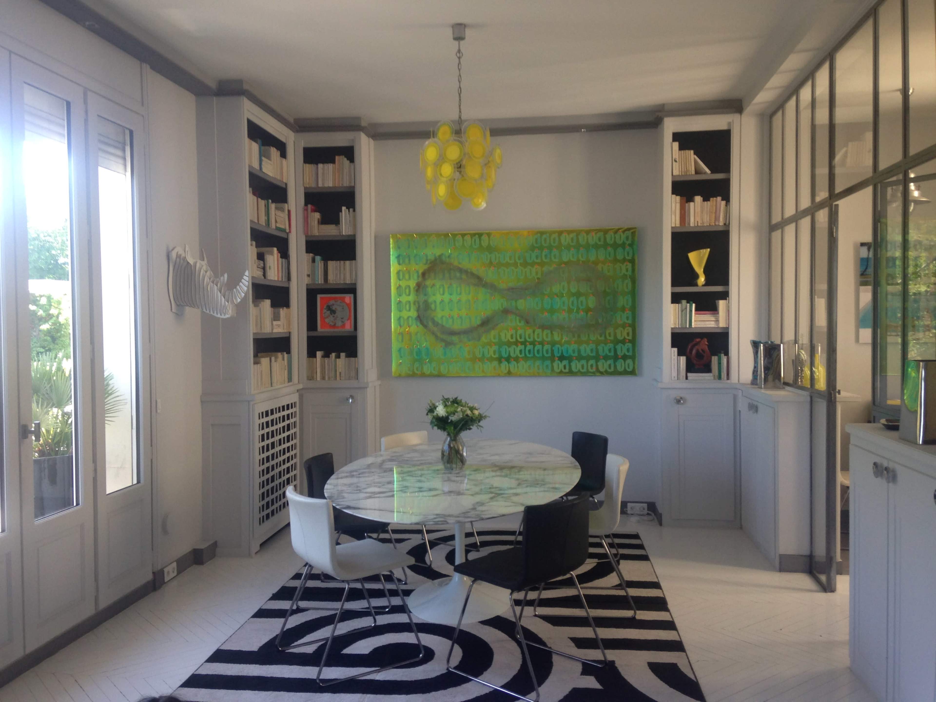 A modern dining room features a round marble table surrounded by black and white chairs, with a bright yellow chandelier hanging above and a large abstract painting on the wall.