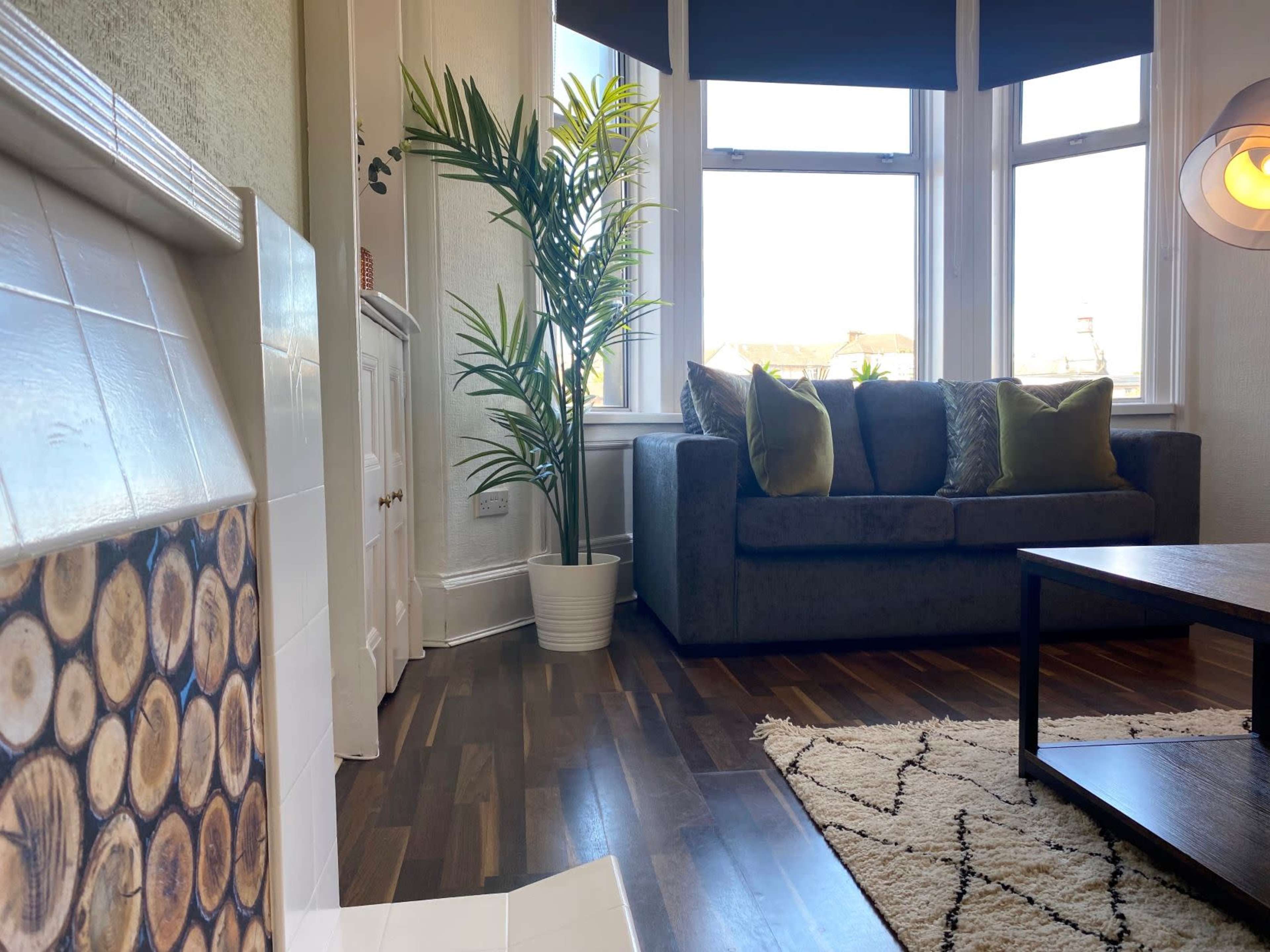 A living room with a dark sofa, a patterned rug, a coffee table, and a large window letting in natural light, complemented by a potted plant in the corner.