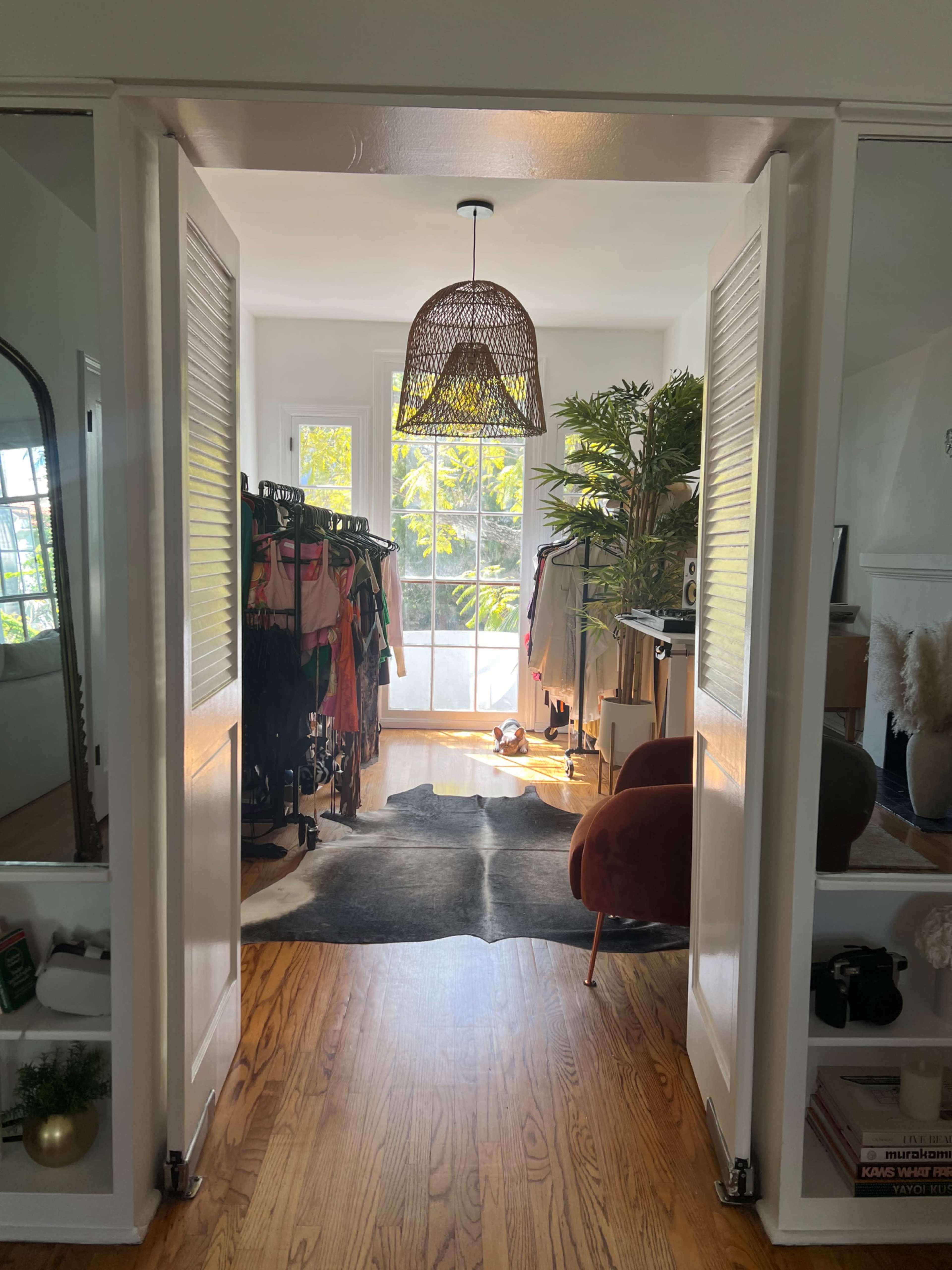 A stylish room is visible through an open door, featuring a woven light fixture, a clothing rack, a large window with natural light, and a cozy seating area.