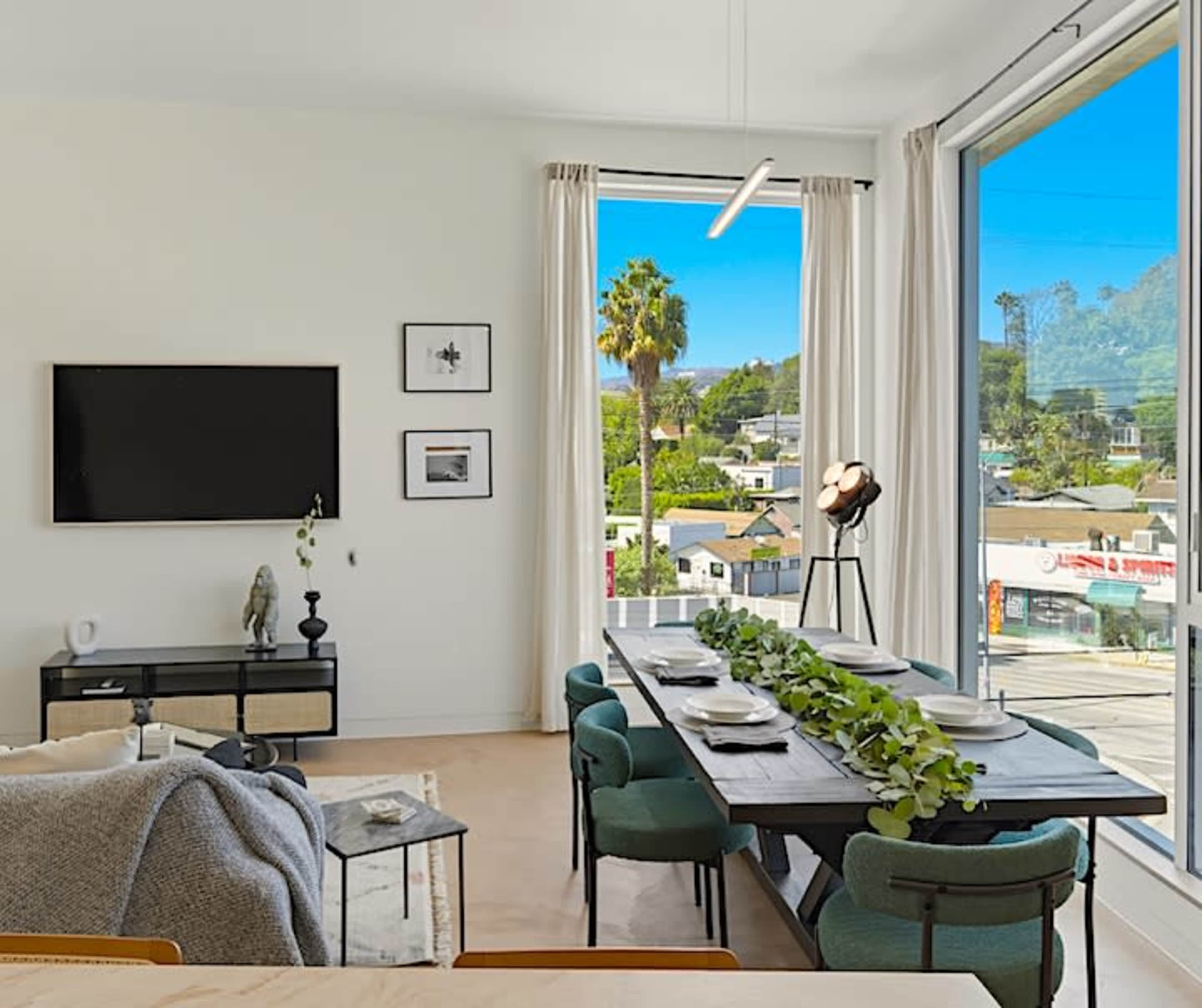A modern dining area features a long table set for a meal, with large windows providing a view of a palm tree and neighborhood outside.