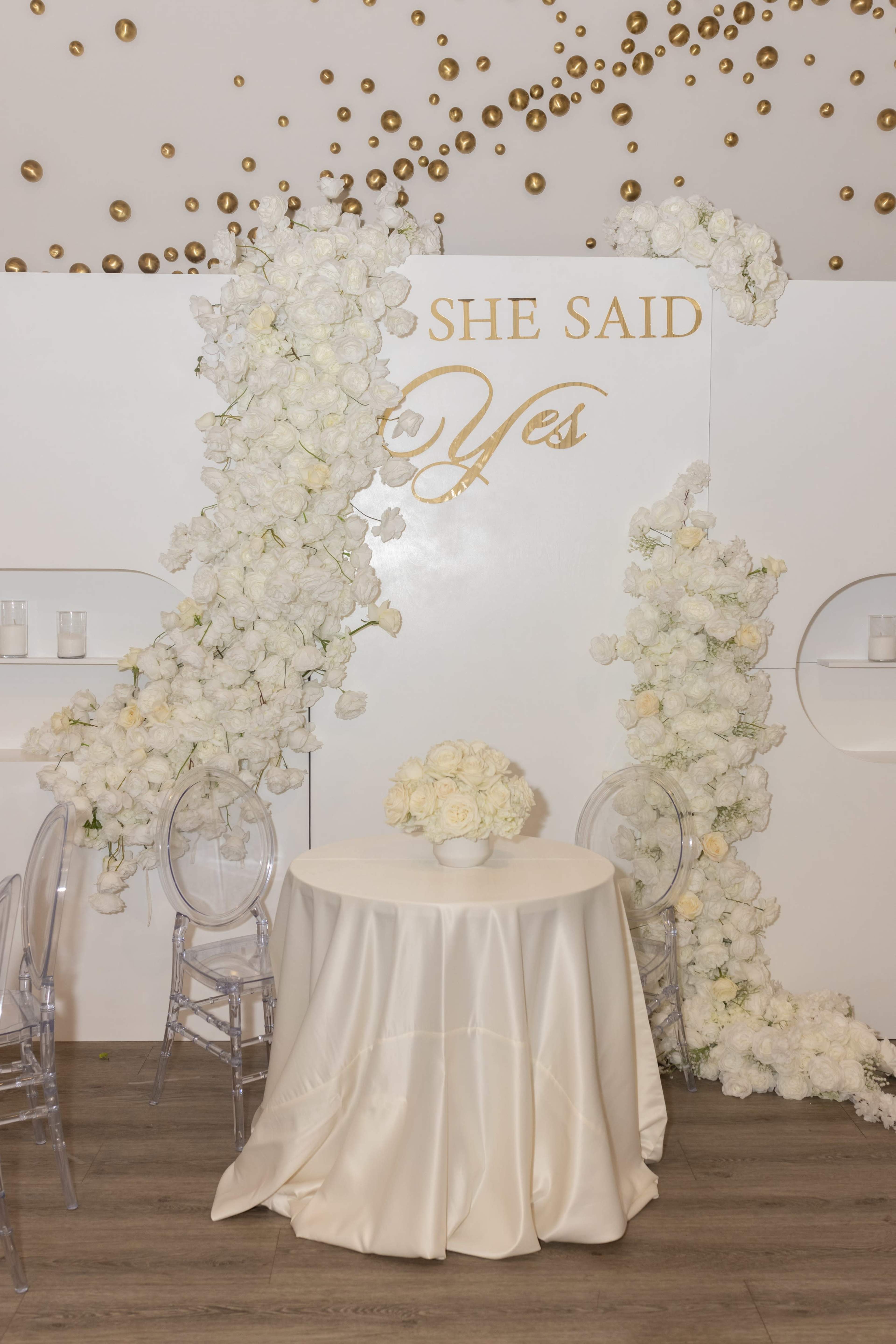 A table draped in a cream-colored cloth is set against a white wall adorned with artificial white flowers and the phrase "SHE SAID Yes" in gold lettering.
