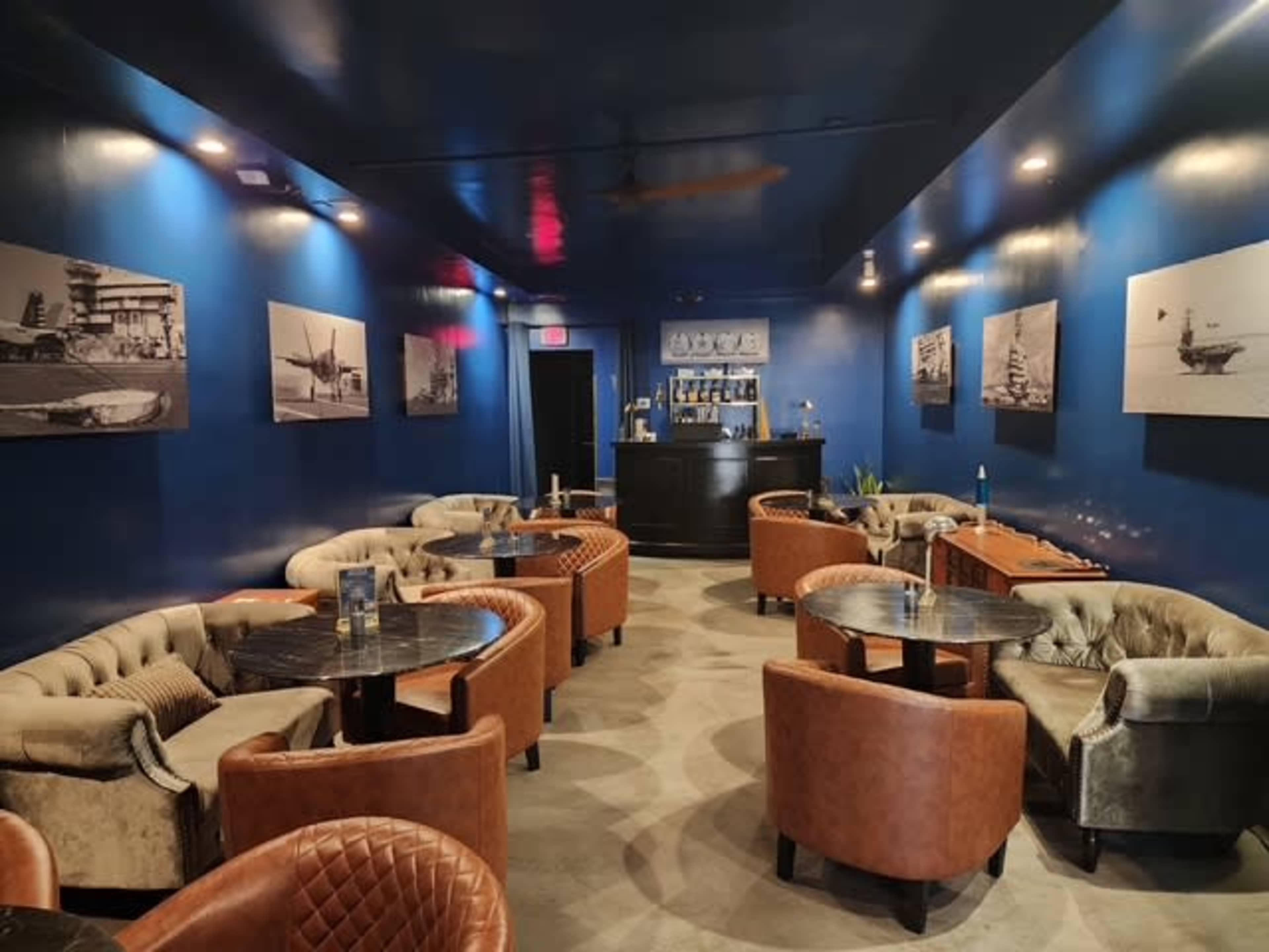 The image shows a modern lounge with dark blue walls, leather seating arrangements, and black tables, featuring black-and-white photographs on the walls.