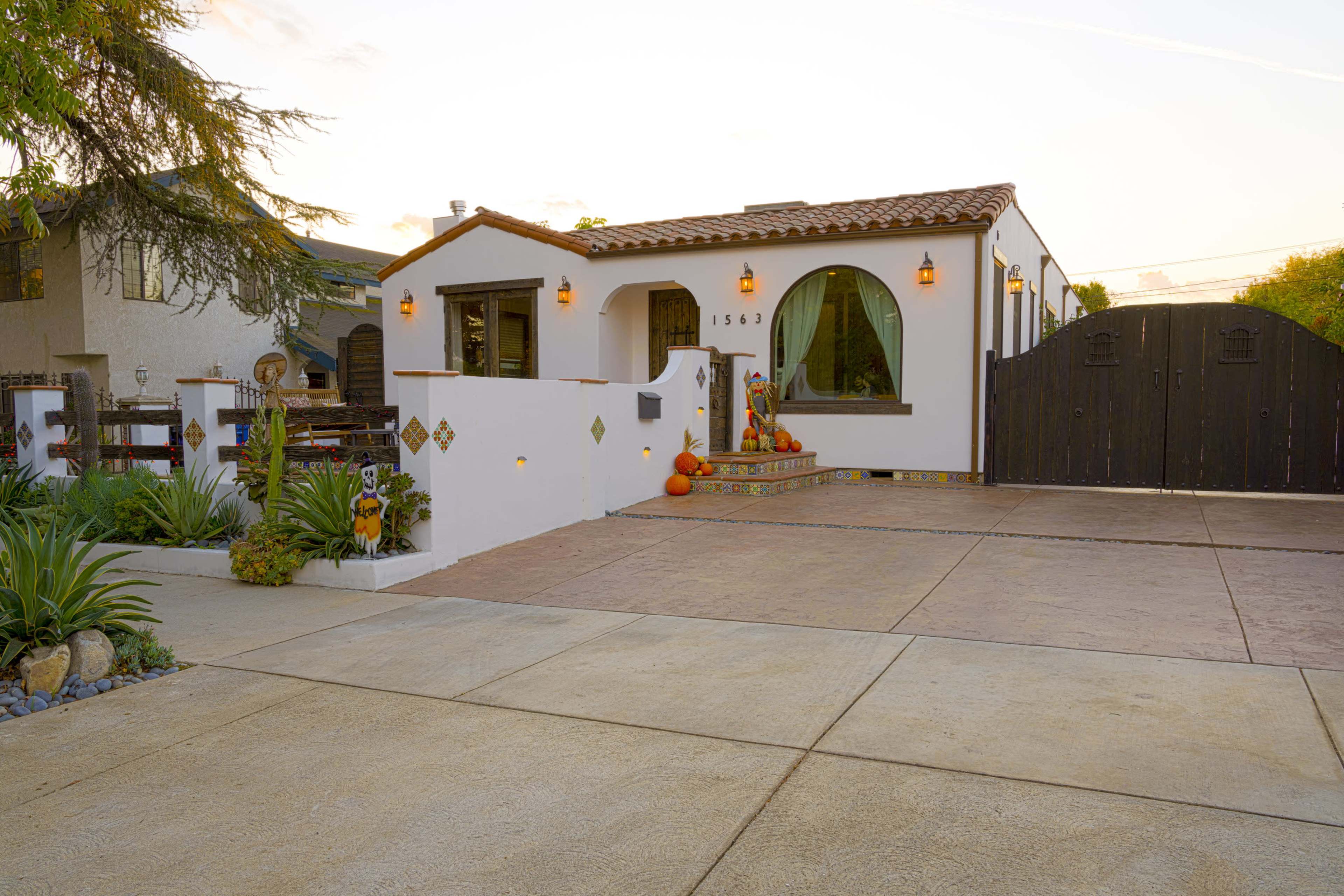 A single-story house with a tiled roof, decorative lights, and pumpkin decorations is set behind a black wooden gate and surrounded by landscaped greenery.