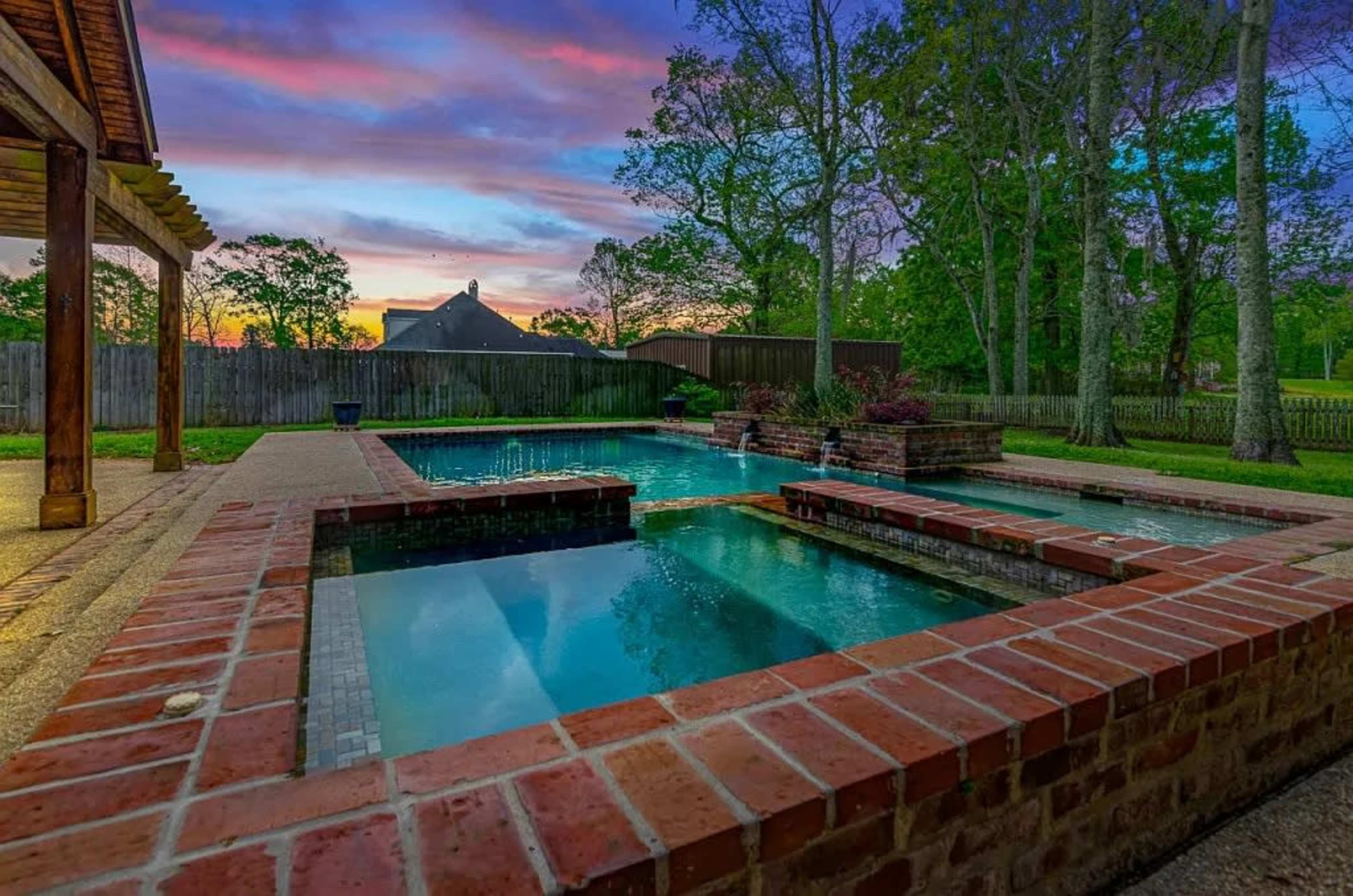 A brick patio surrounds a pool and hot tub, with trees and a colorful sunset visible in the background.
