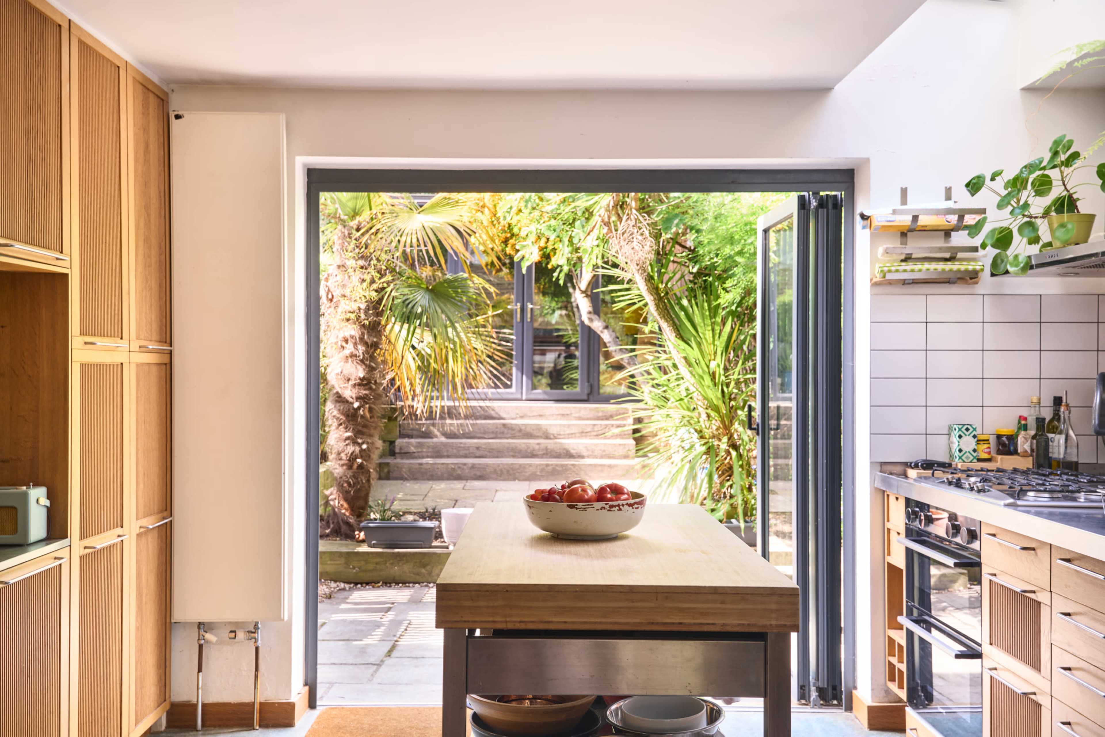 The image shows a modern kitchen with an island in the center and a view of a garden through large open doors.