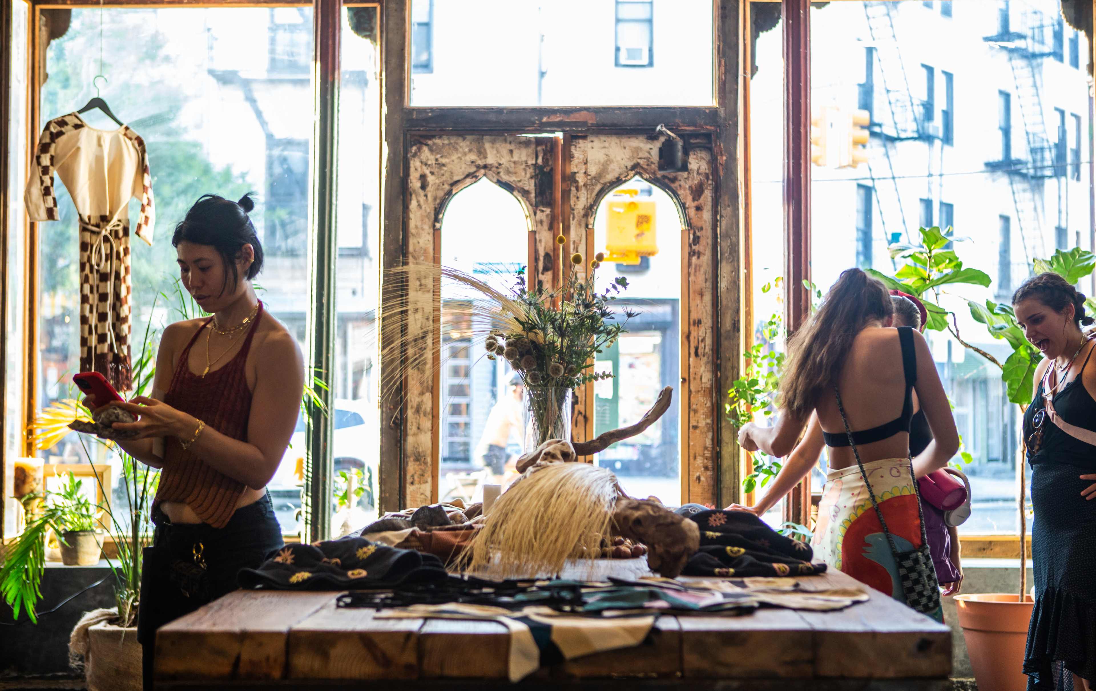A vintage clothing boutique features two women shopping and browsing among colorful garments and displayed accessories in front of large windows.