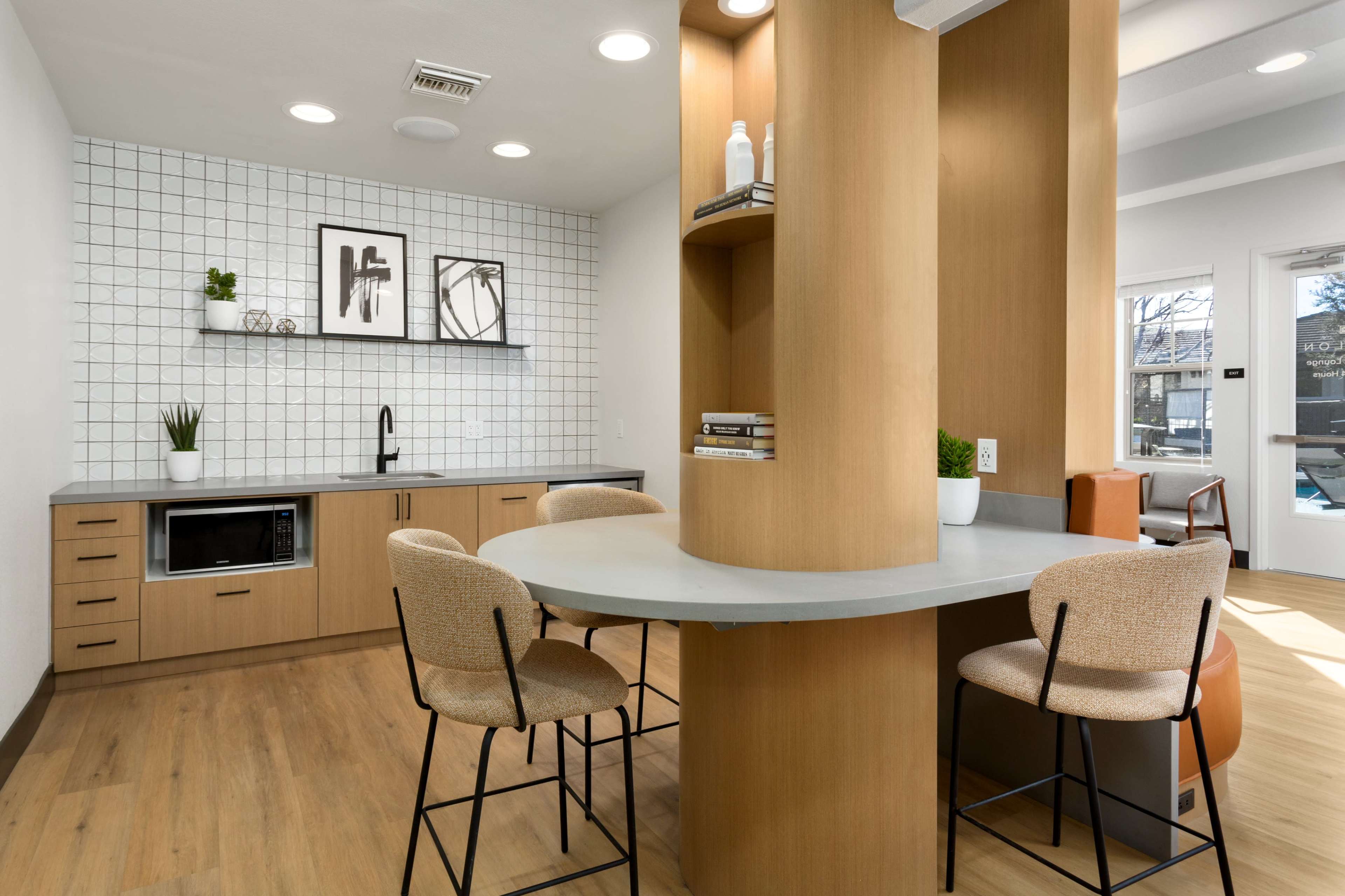 A modern kitchen area with light wood cabinetry, a gray countertop, a microwave, and a small dining table with two bar stools.