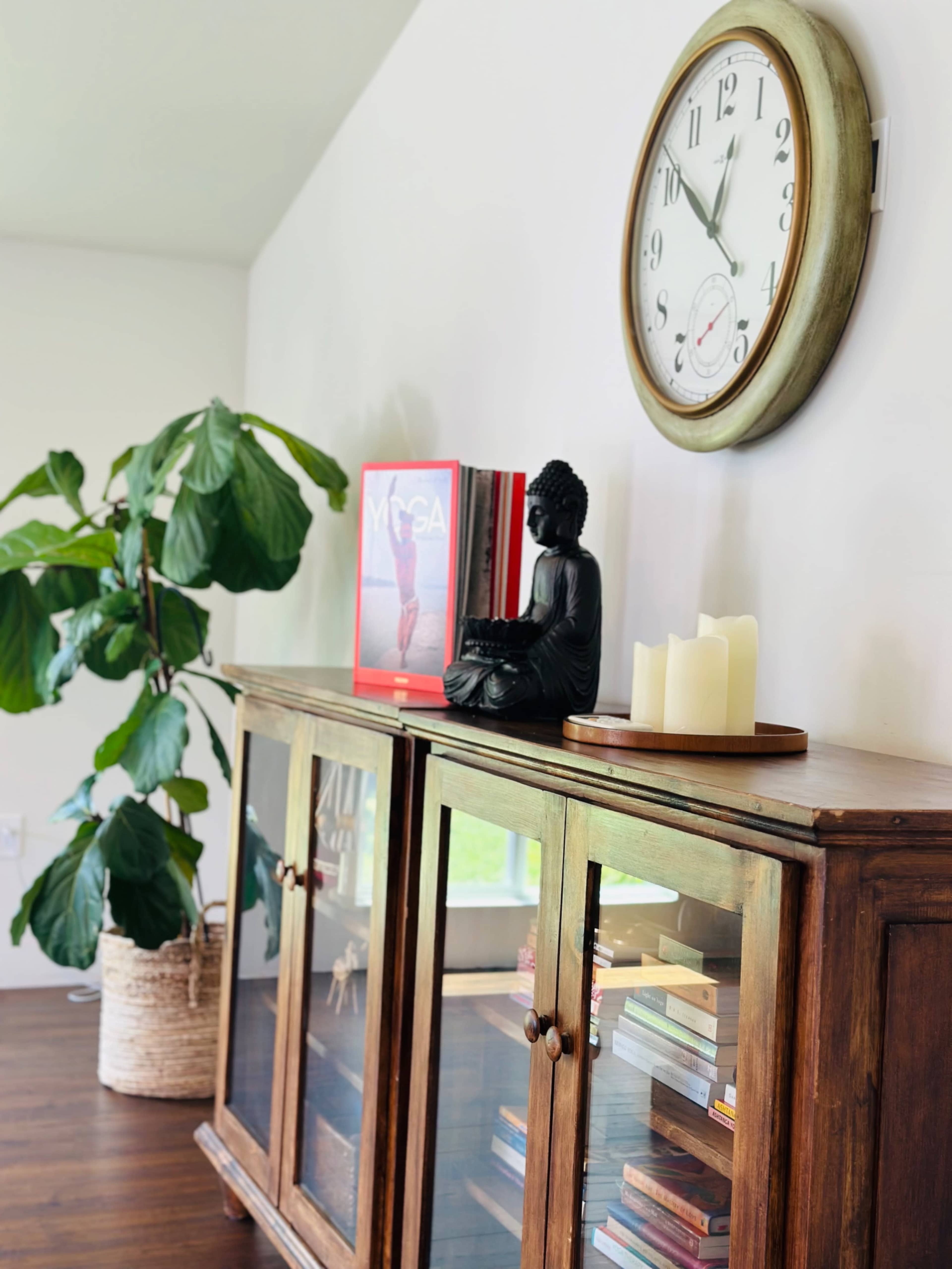 A wooden cabinet with glass doors displays books and a decorative statue, while a large clock and a potted plant are arranged nearby on a wall.