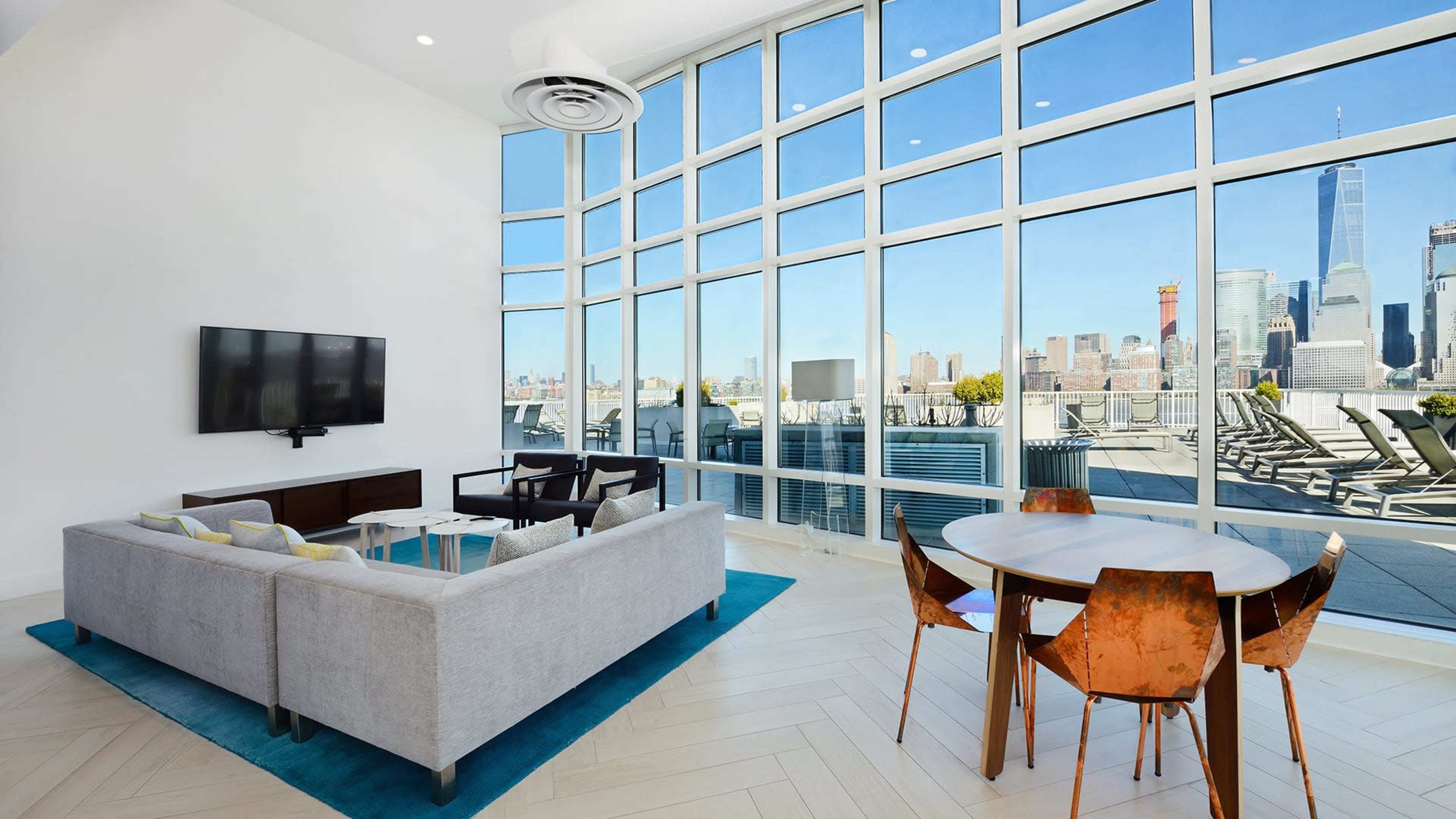 The modern living space features a sectional sofa, a small wooden dining table, and large windows with a view of a city skyline.
