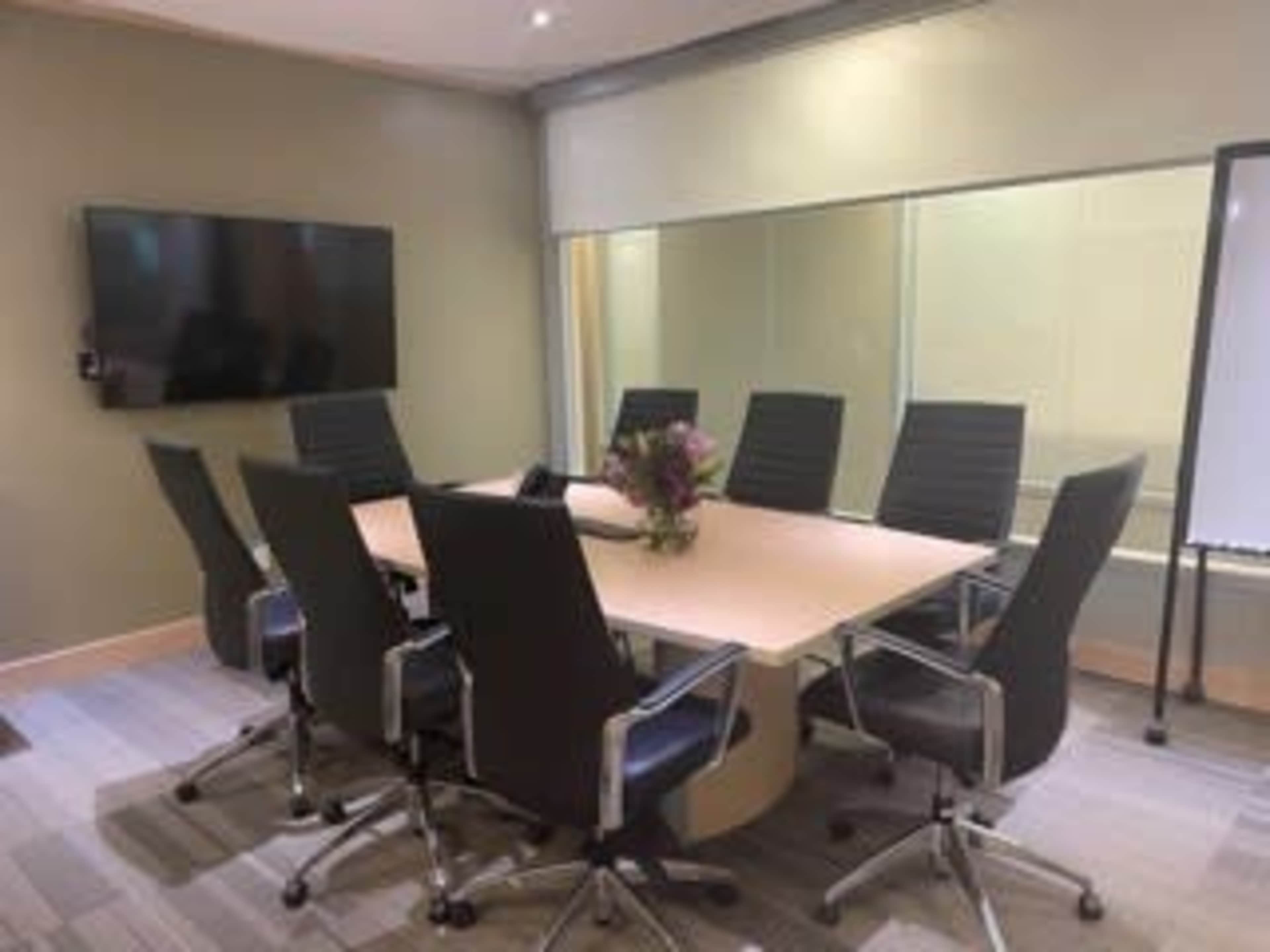 A conference room features a large table surrounded by eight black chairs, with a television and whiteboard on the walls.