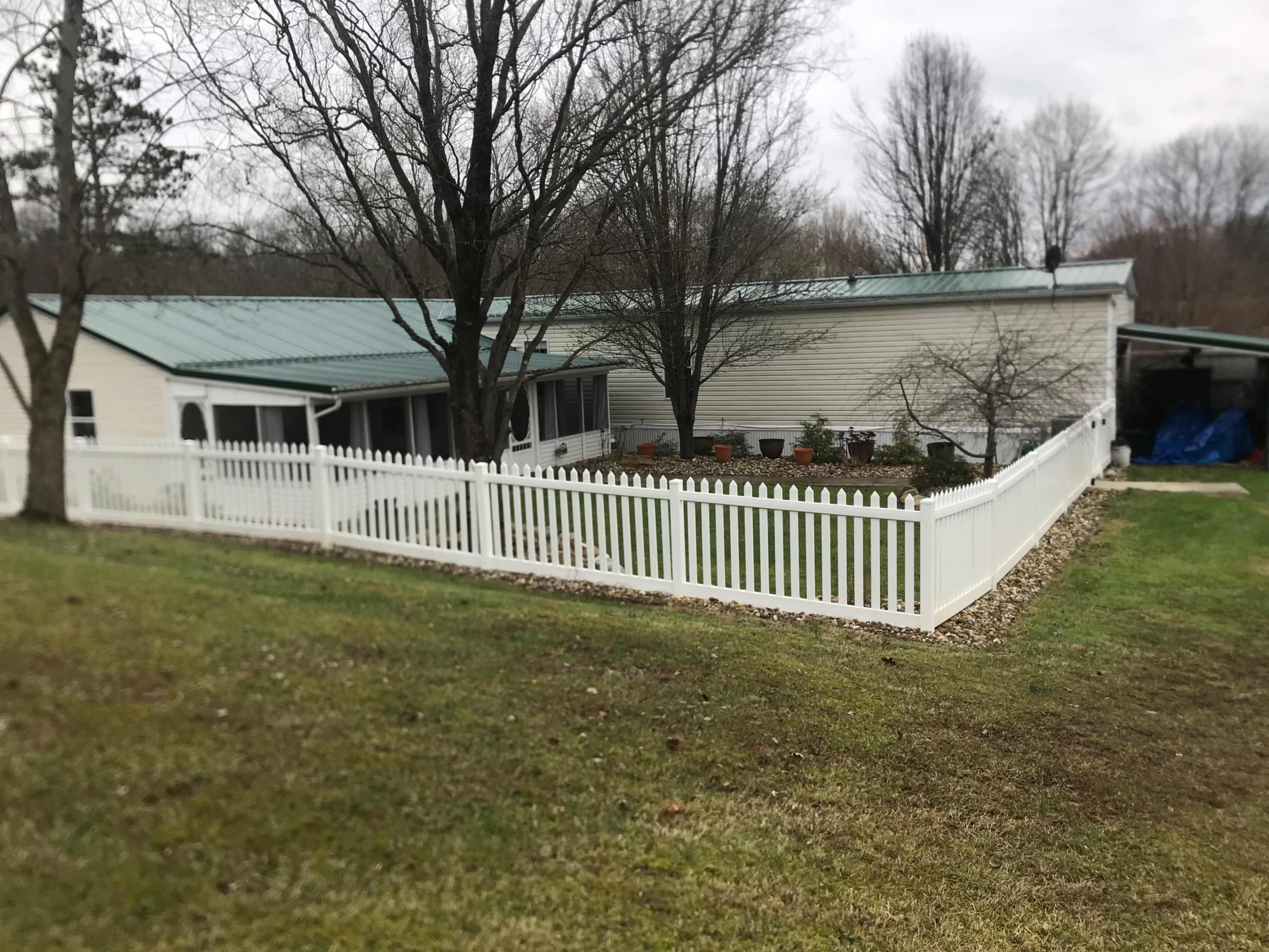 A white picket fence surrounds a single-story house with a metal roof and nearby trees.