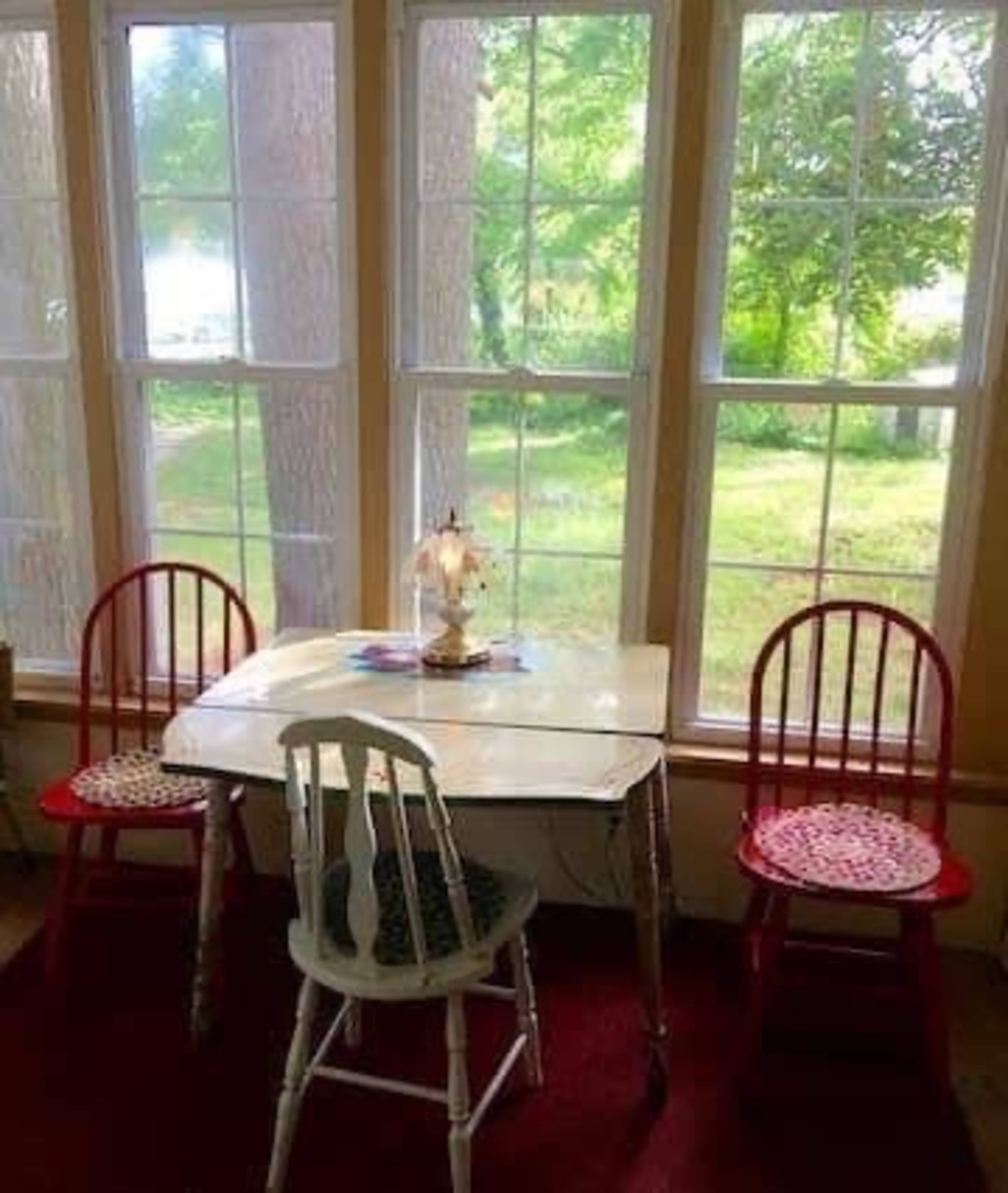 A small table with a lamp sits between three chairs in a sunlit room with large windows overlooking a green landscape.