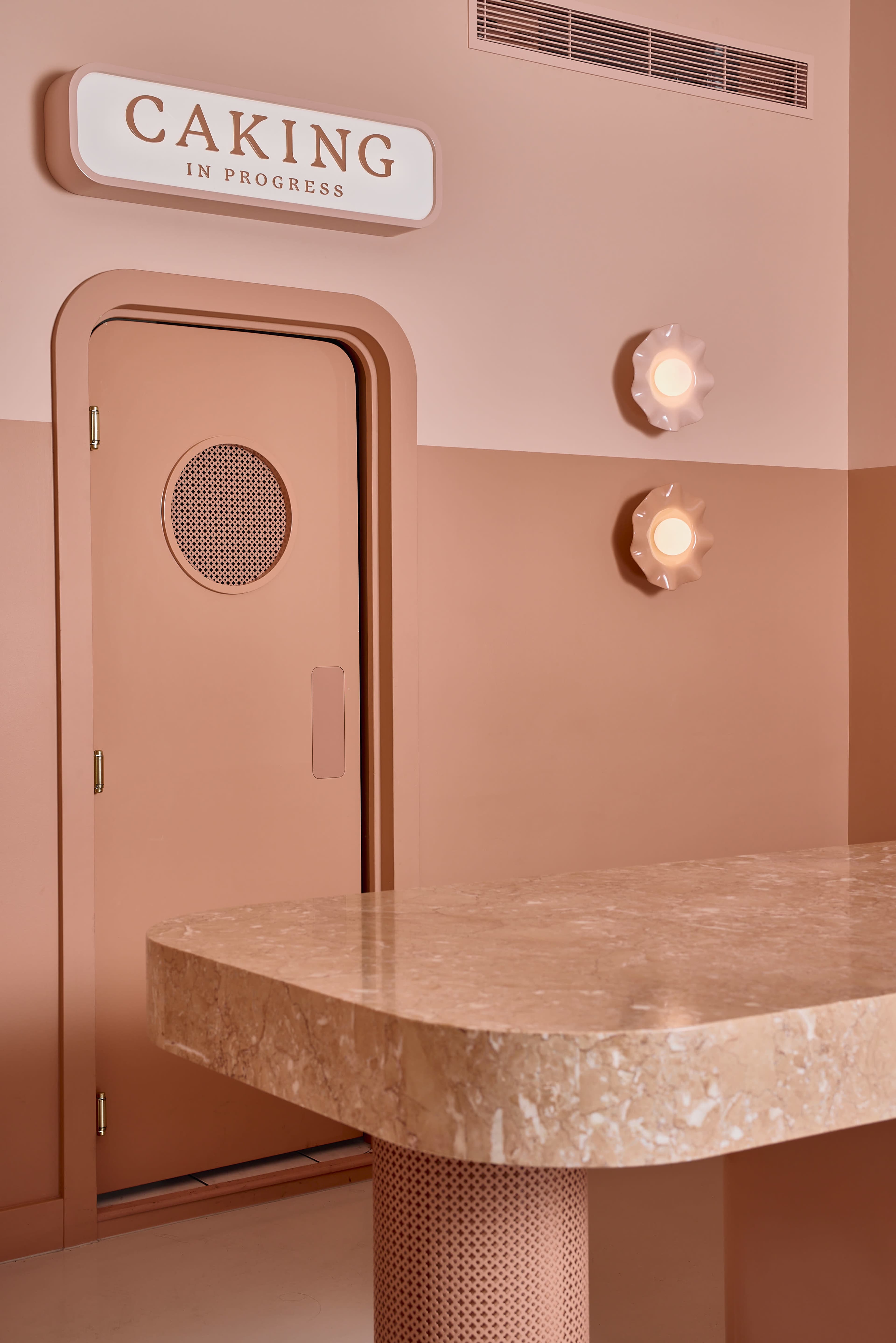The image shows a minimalist interior featuring a pinkish wall, a round table made of marble, and a door with a mesh circular window labeled "CAKING IN PROGRESS."