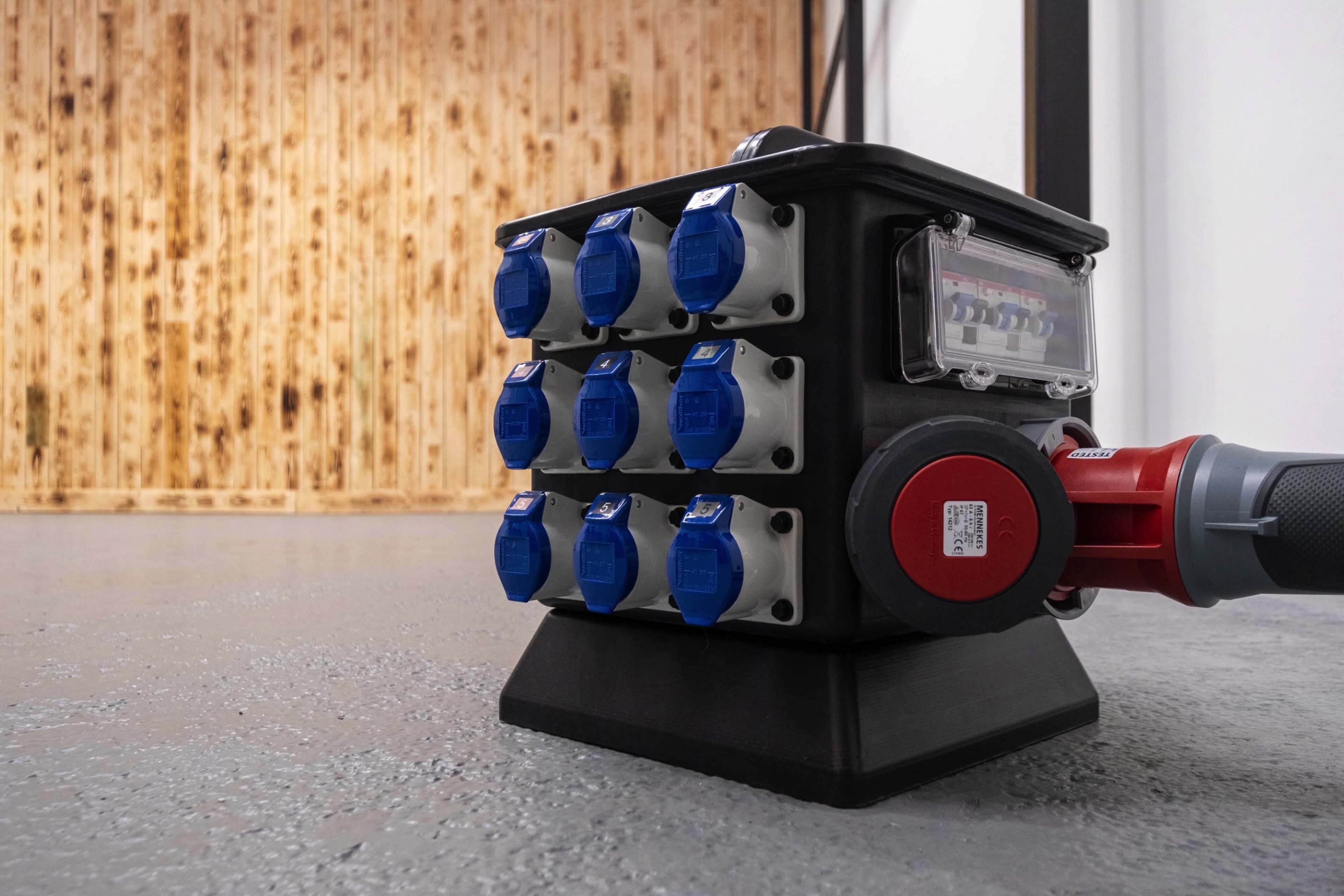 A black electrical distribution box with multiple blue outlets is positioned on a flat surface in an industrial setting.