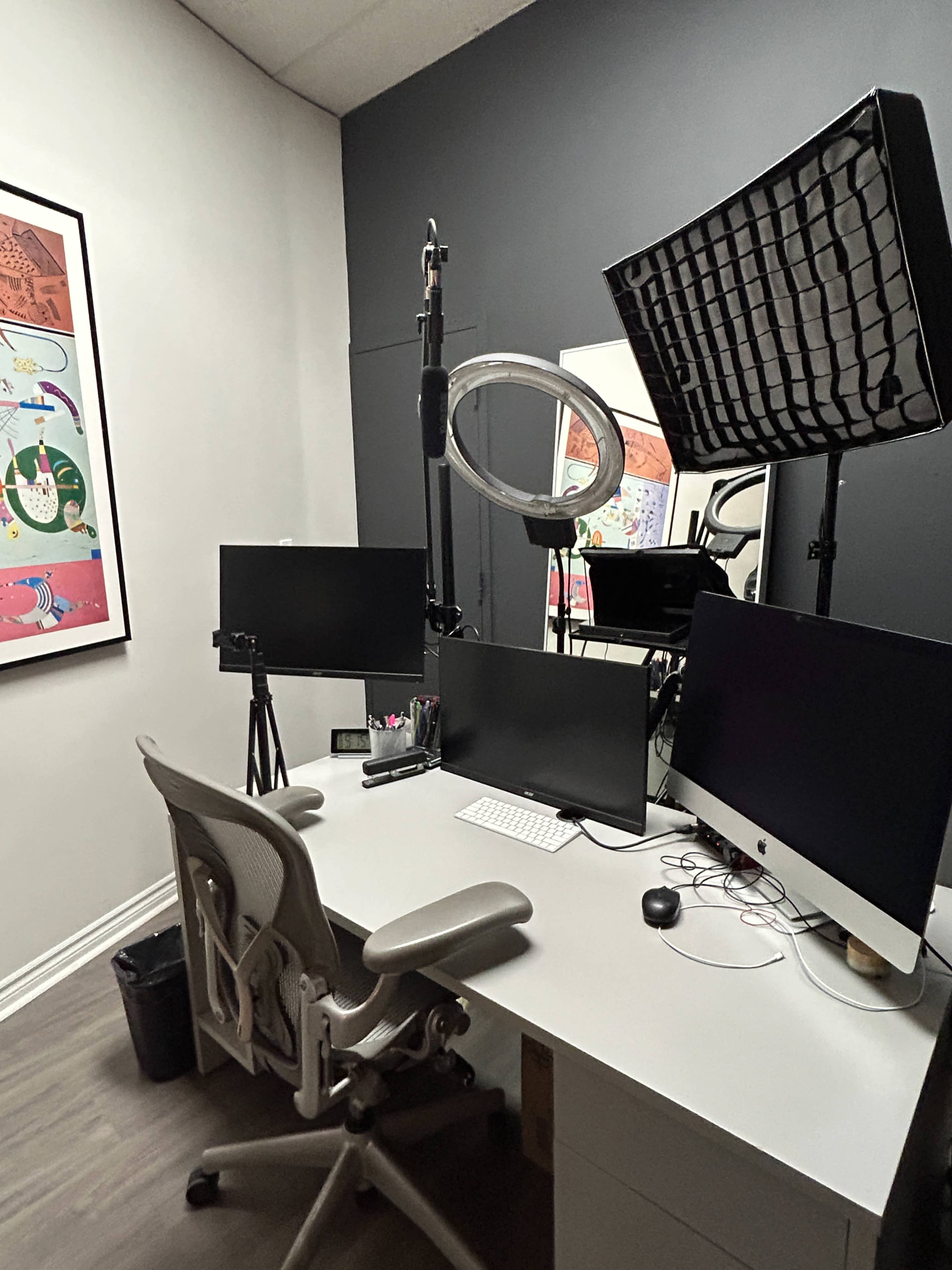 The image shows a modern office space featuring a desk with two computer monitors, a keyboard, a chair, and professional lighting equipment mounted on stands.