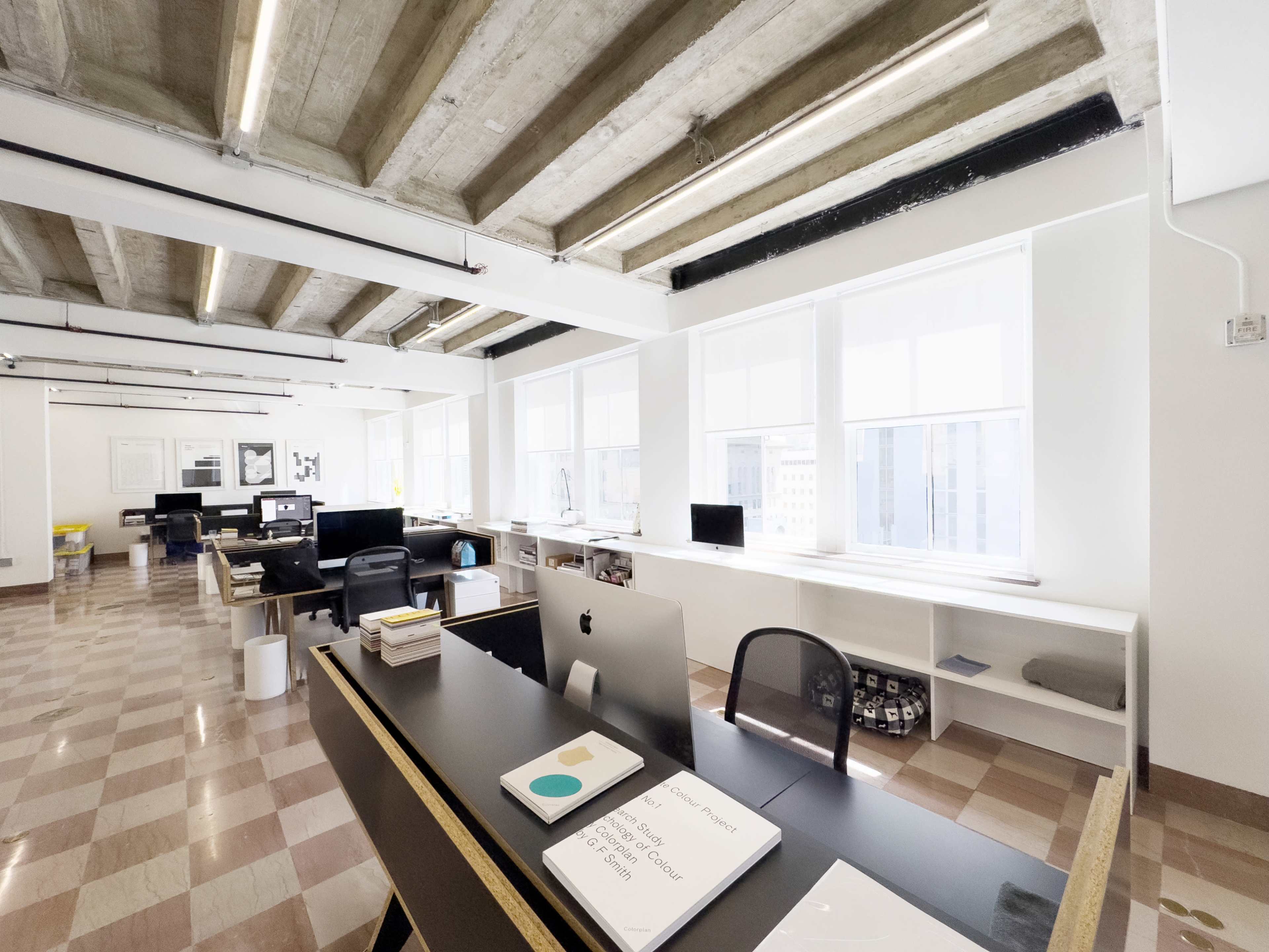 A modern office space features large windows, a sleek black desk with an iMac, and organized workstations with computer screens and minimalist decor.