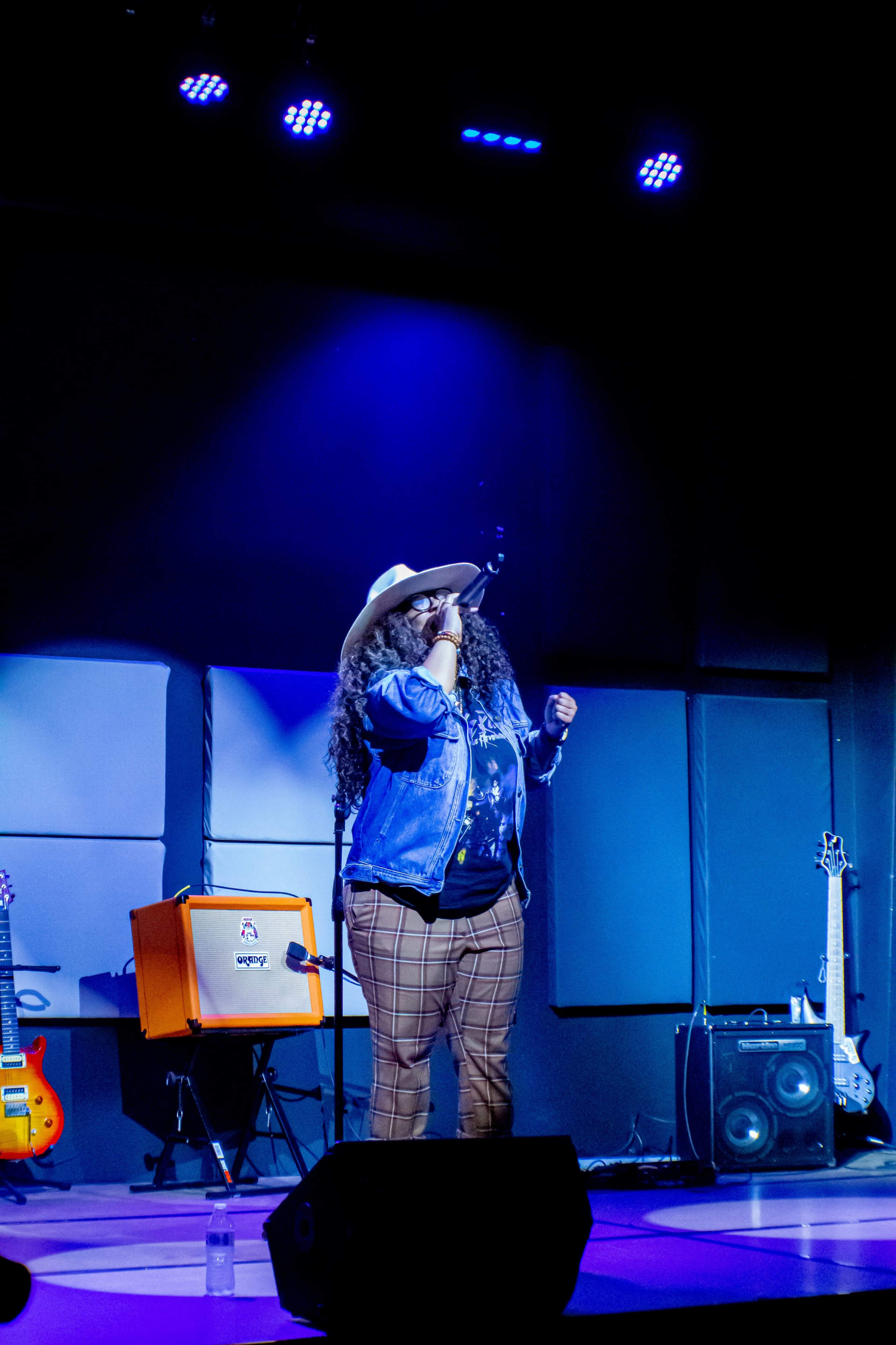 A performer sings on stage while wearing a hat and denim jacket, with musical instruments and equipment visible in the background.