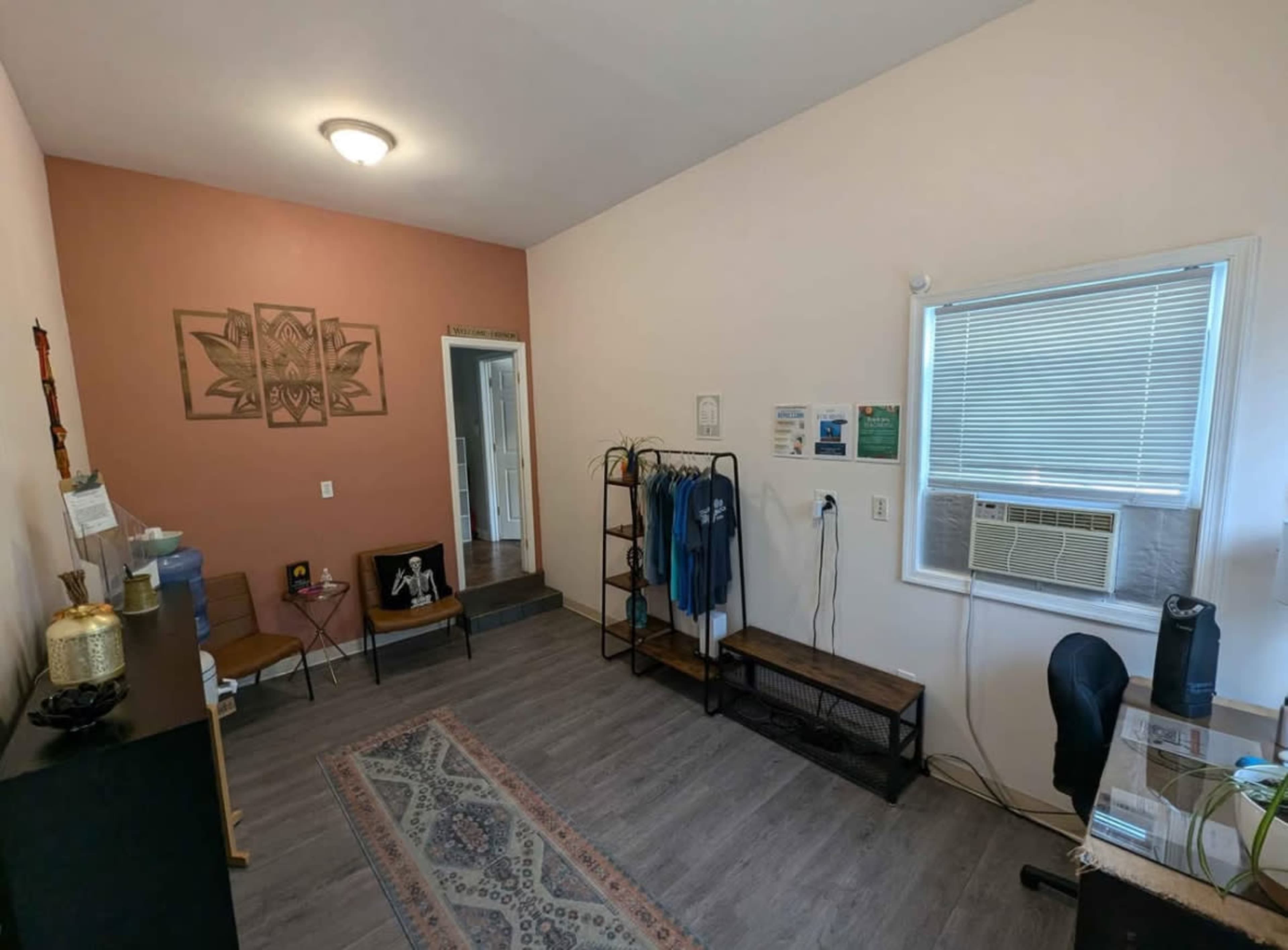 A small, well-lit room features a desk, seating area, and shelves with clothing, alongside a door leading to another space.