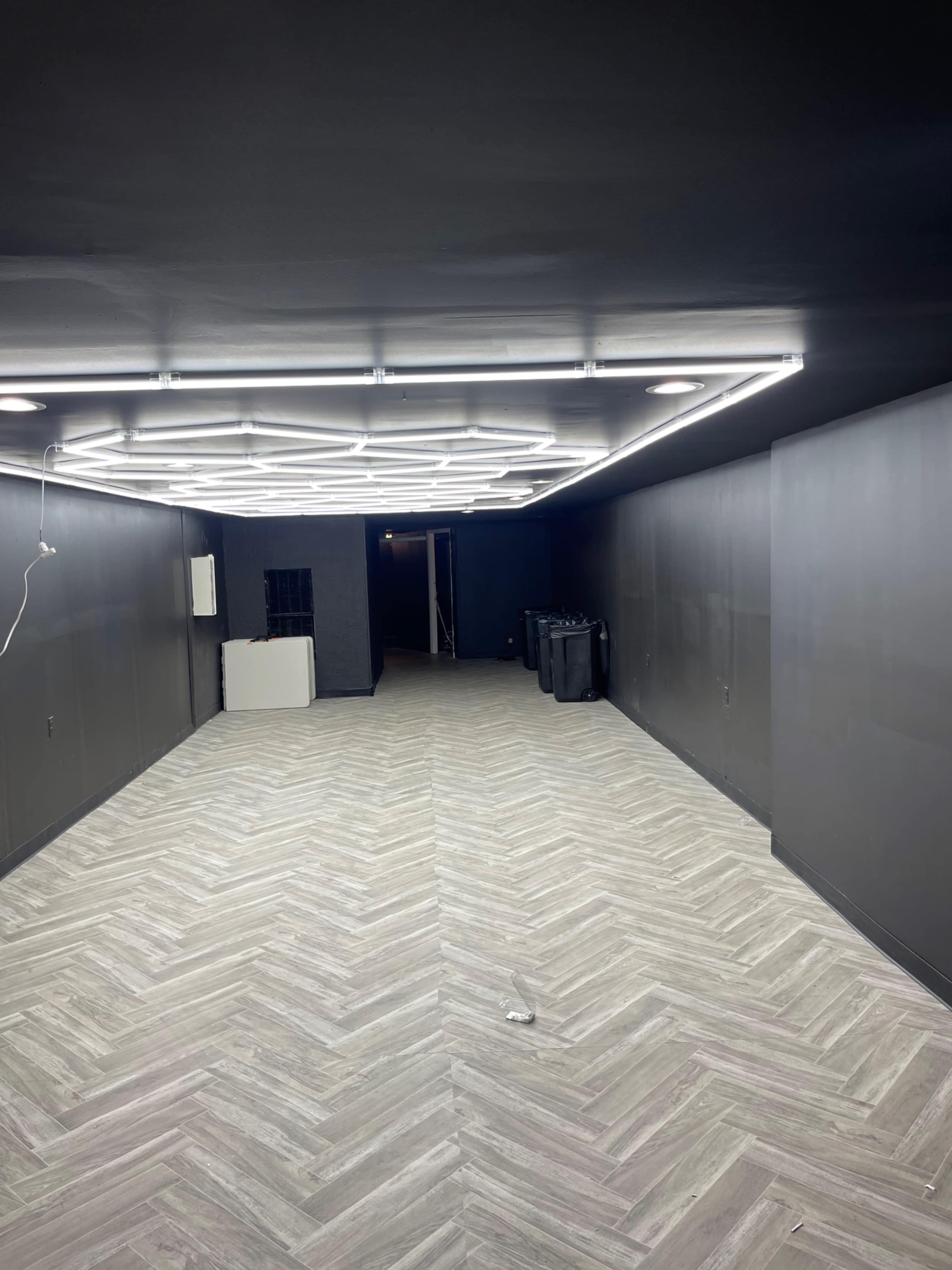 An empty room with dark walls, a herringbone-patterned floor, and bright overhead lighting.