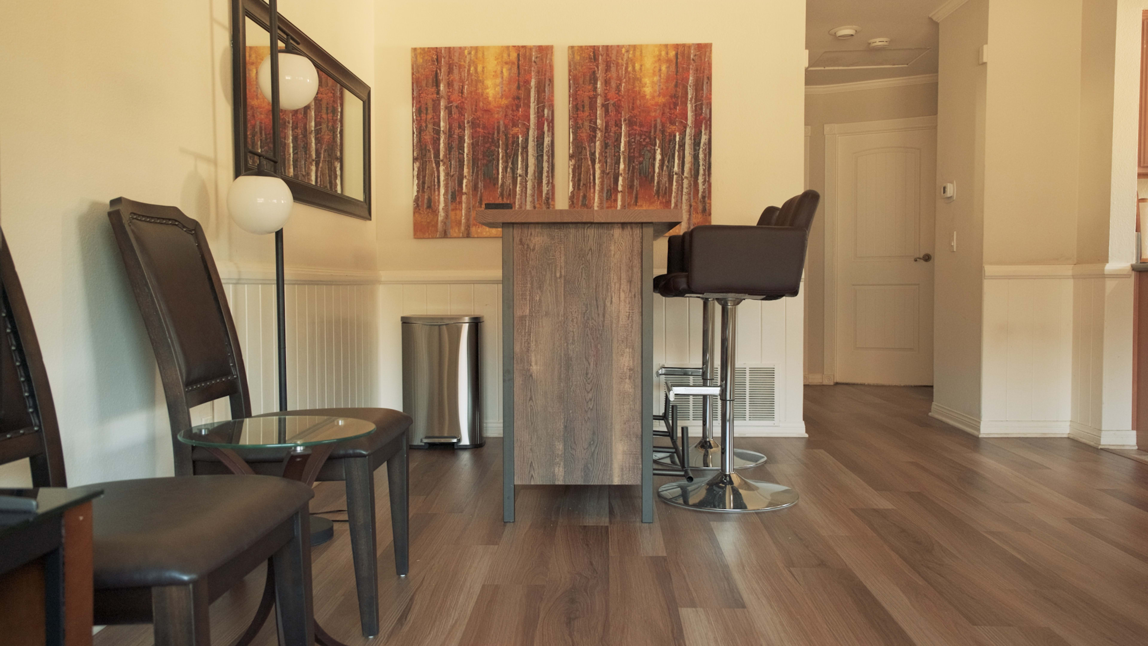A modern dining area with a wooden bar table and high stools, framed by artwork featuring autumn trees and a trash can in the corner.