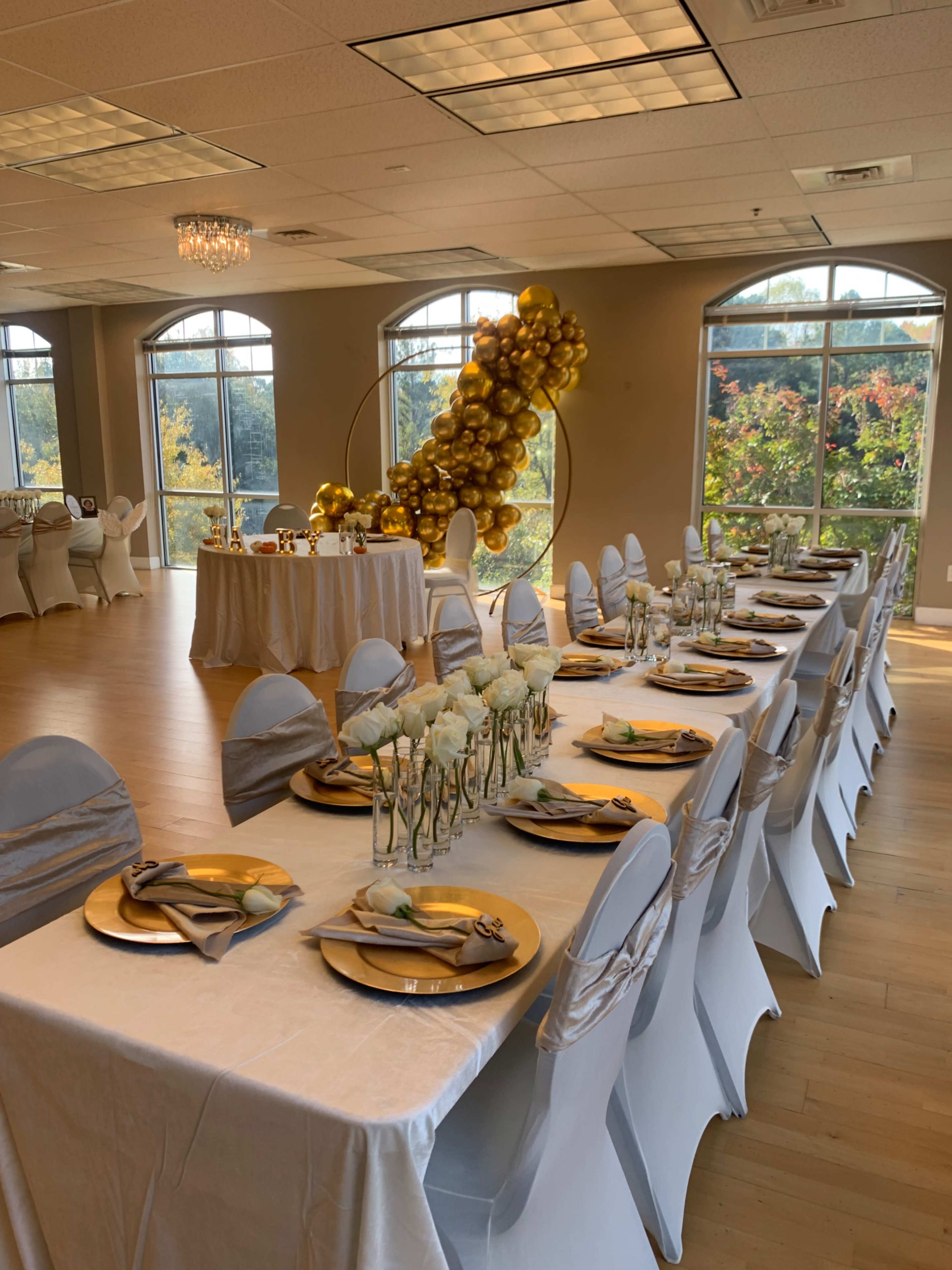 A long banquet table is elegantly set with gold plates and flowers, surrounded by white chairs adorned with gold accents, in a well-lit event space featuring large windows.