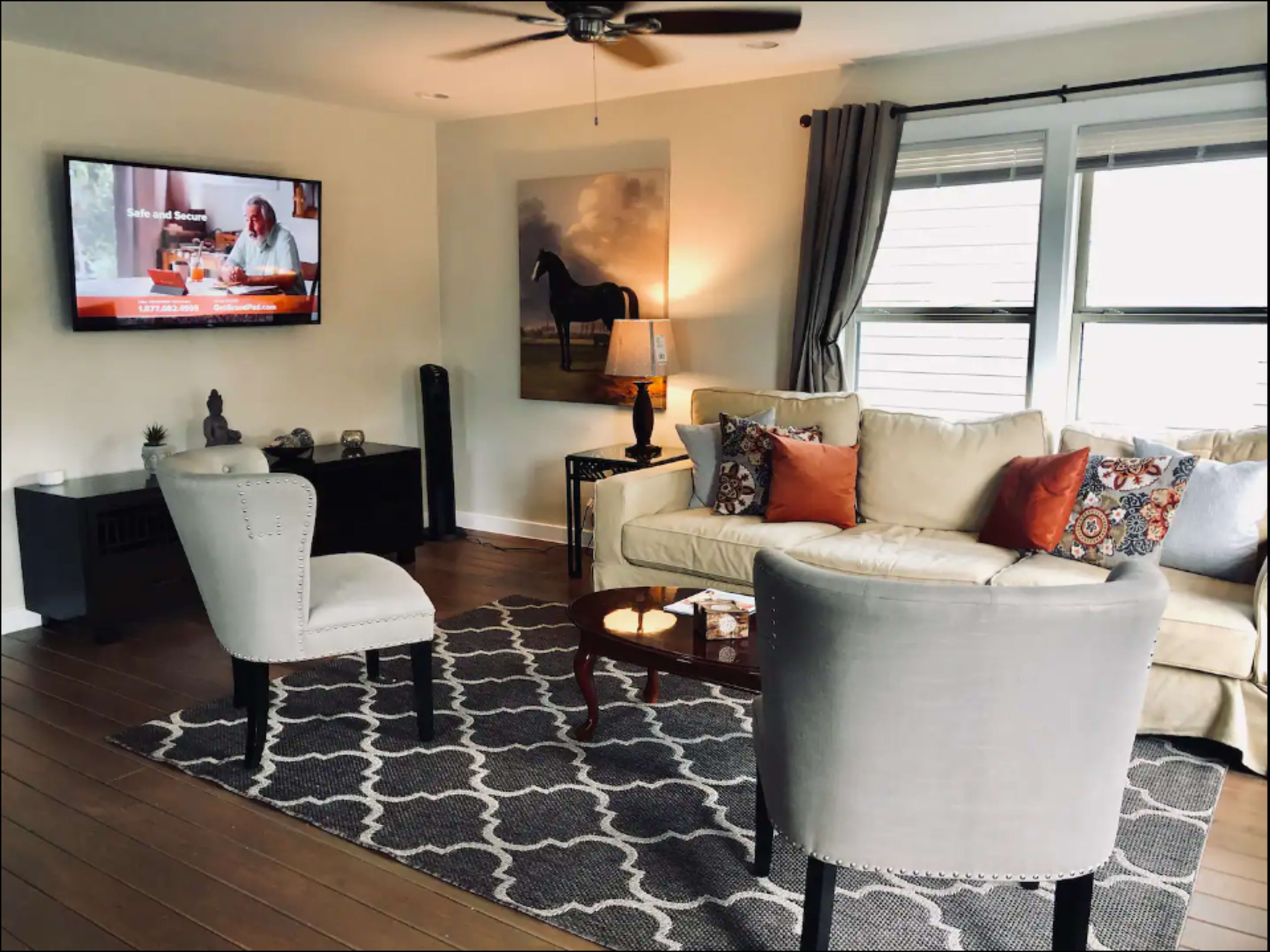 The living room features a light-colored sofa with decorative pillows, two gray chairs, a wooden coffee table, and a television mounted on the wall.