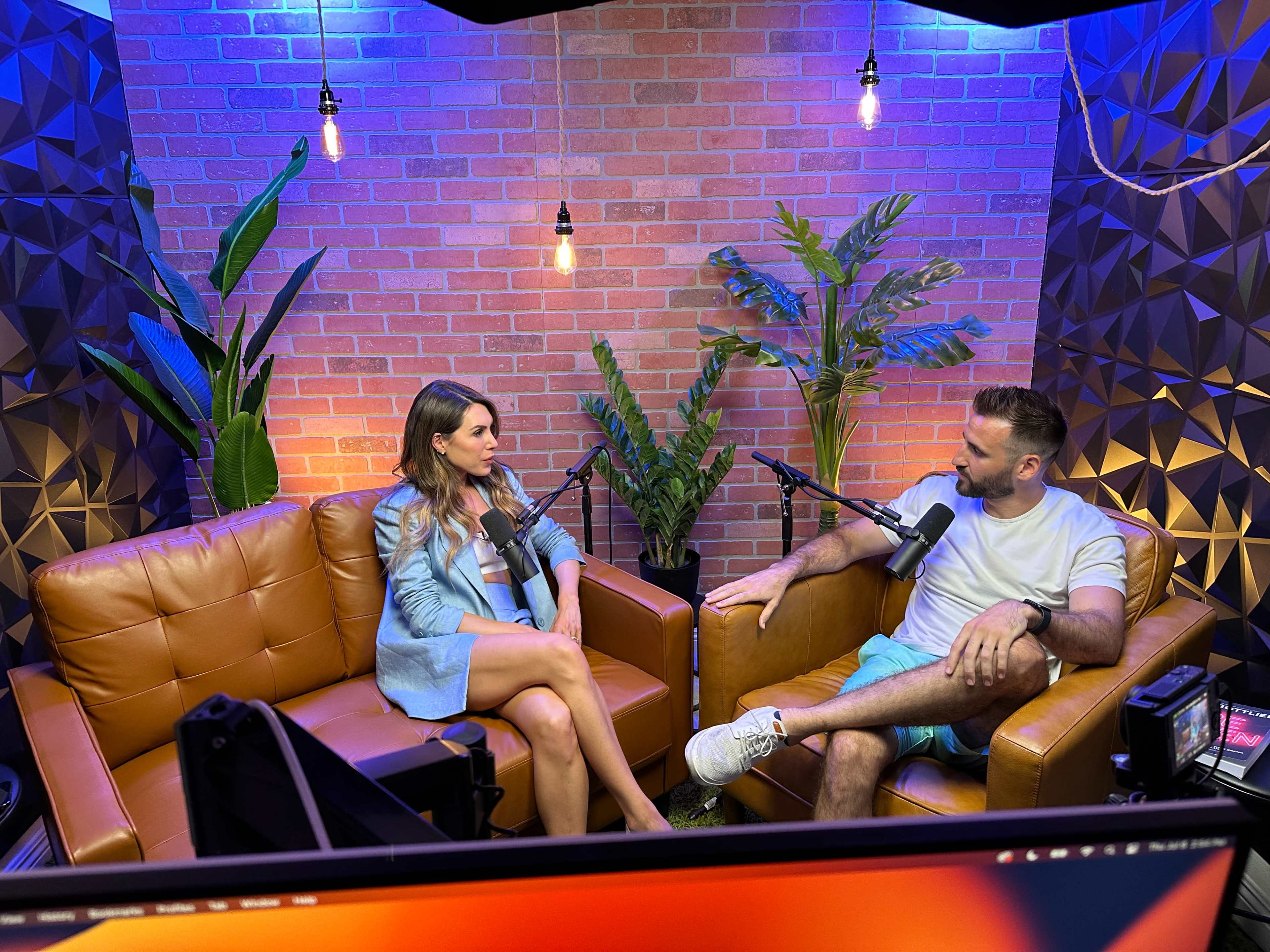 A woman and a man sit on brown couches in a well-lit studio, engaged in conversation with microphones in front of them, surrounded by plants and decorative brick walls.