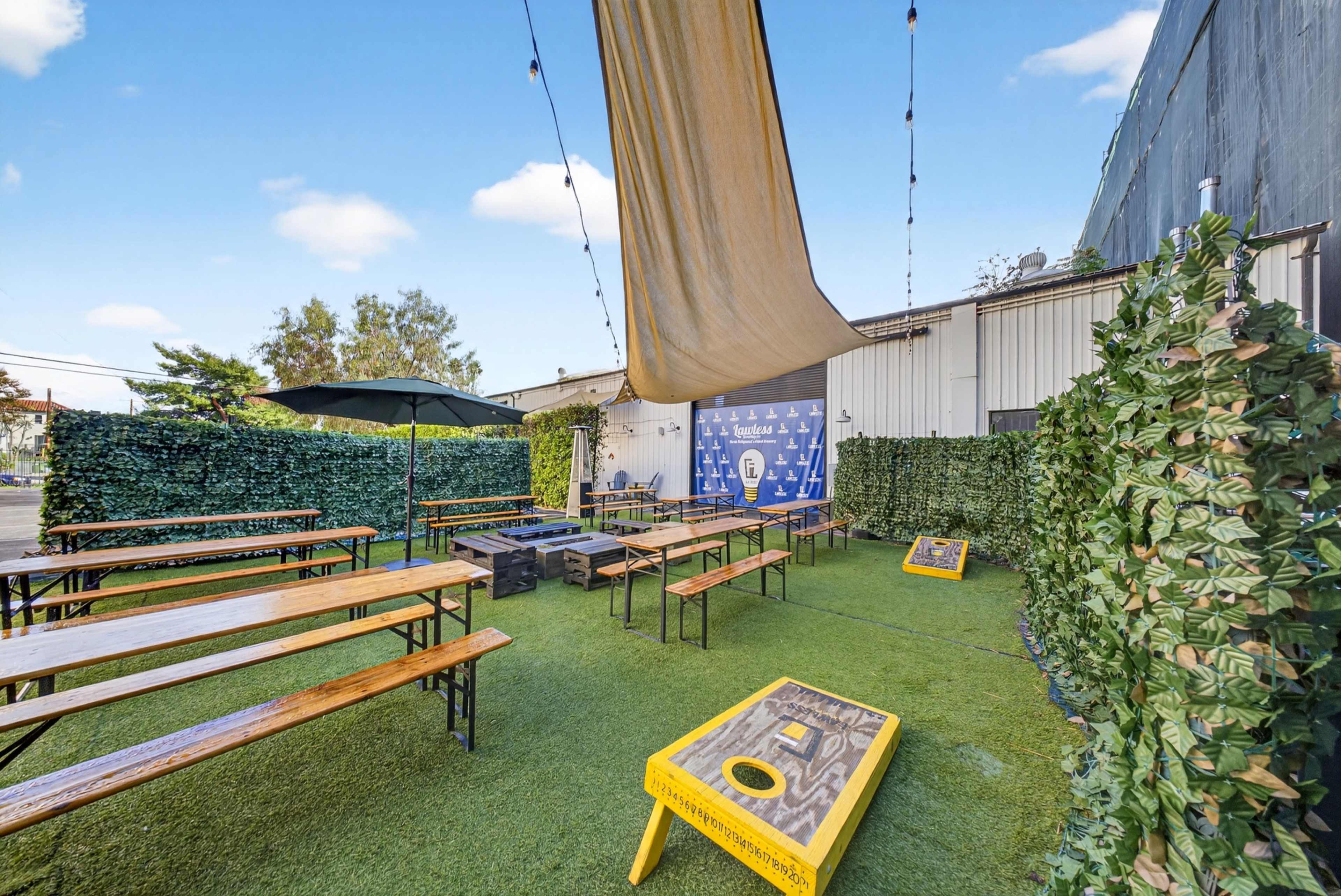 The image shows an outdoor seating area with wooden benches, a large fabric canopy, and cornhole games set up on a grassy surface, surrounded by lush greenery.
