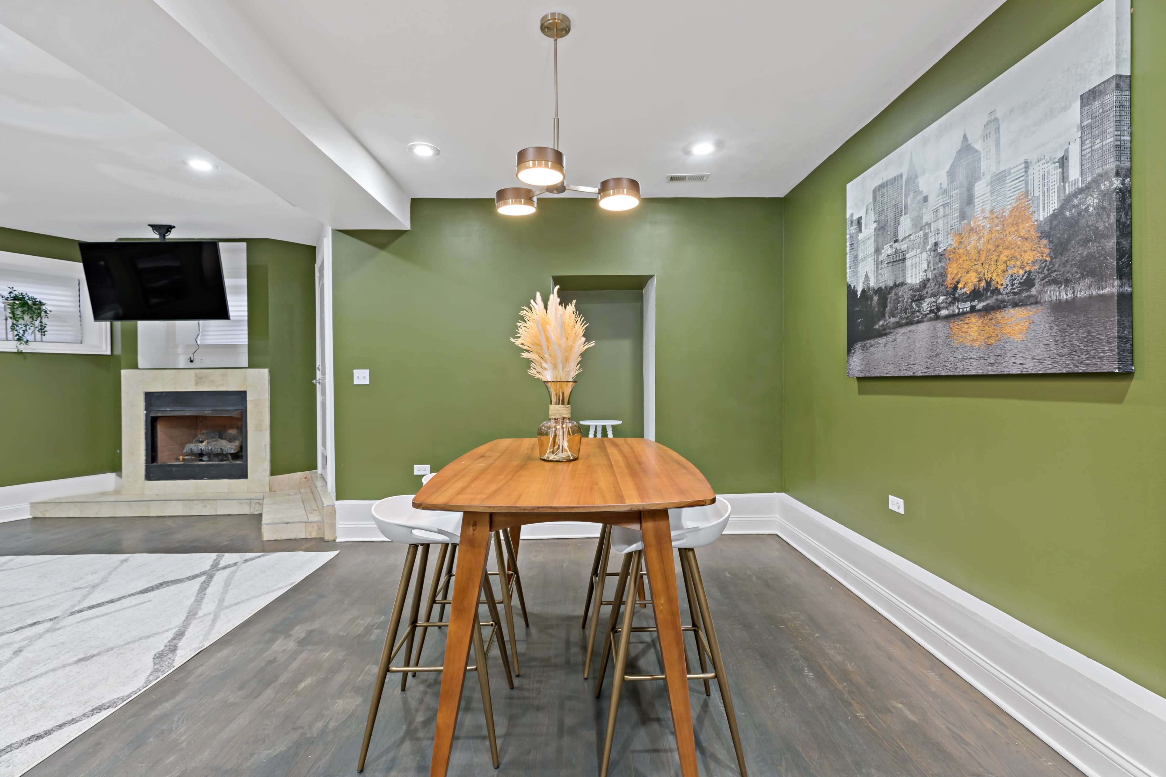 A dining area features a wooden table with two chairs, a wall-mounted television, and a fireplace in a room painted green with a framed cityscape on the wall.