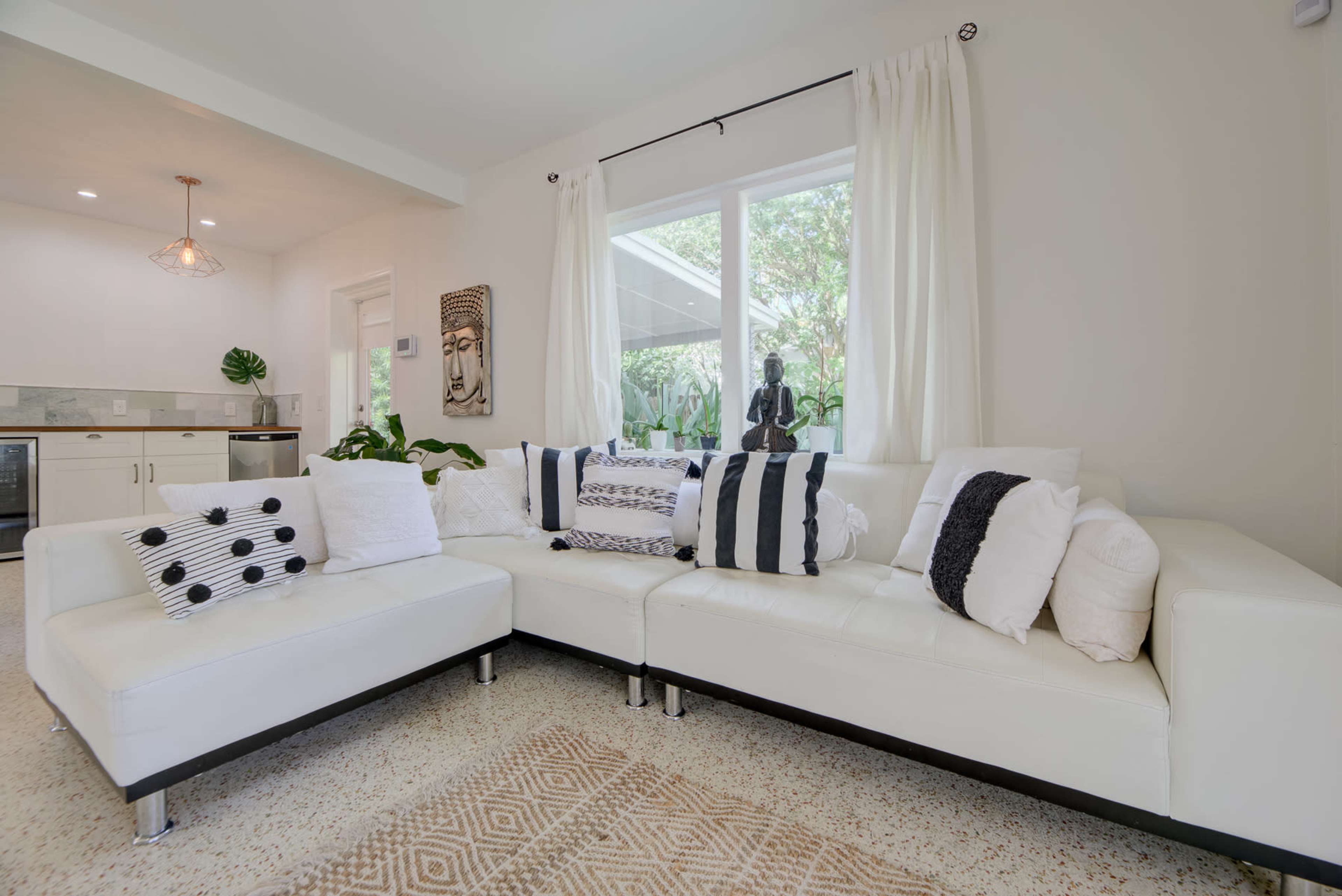 The image shows a spacious living room featuring a white sectional sofa with decorative pillows, a textured area rug, and natural light streaming through a large window.