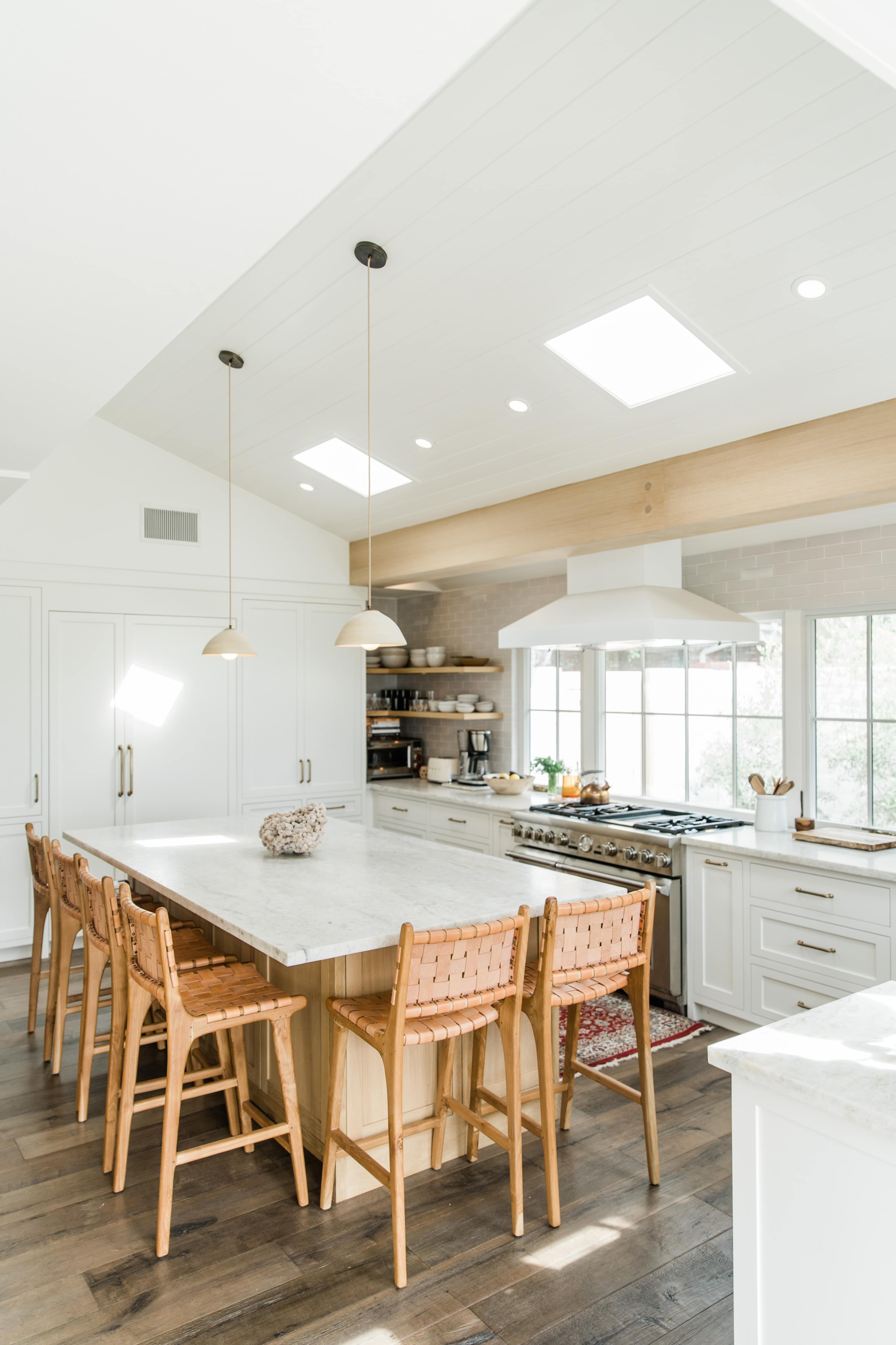 A modern kitchen features a large marble island with woven chairs, stainless steel appliances, and plenty of natural light from skylights and windows.