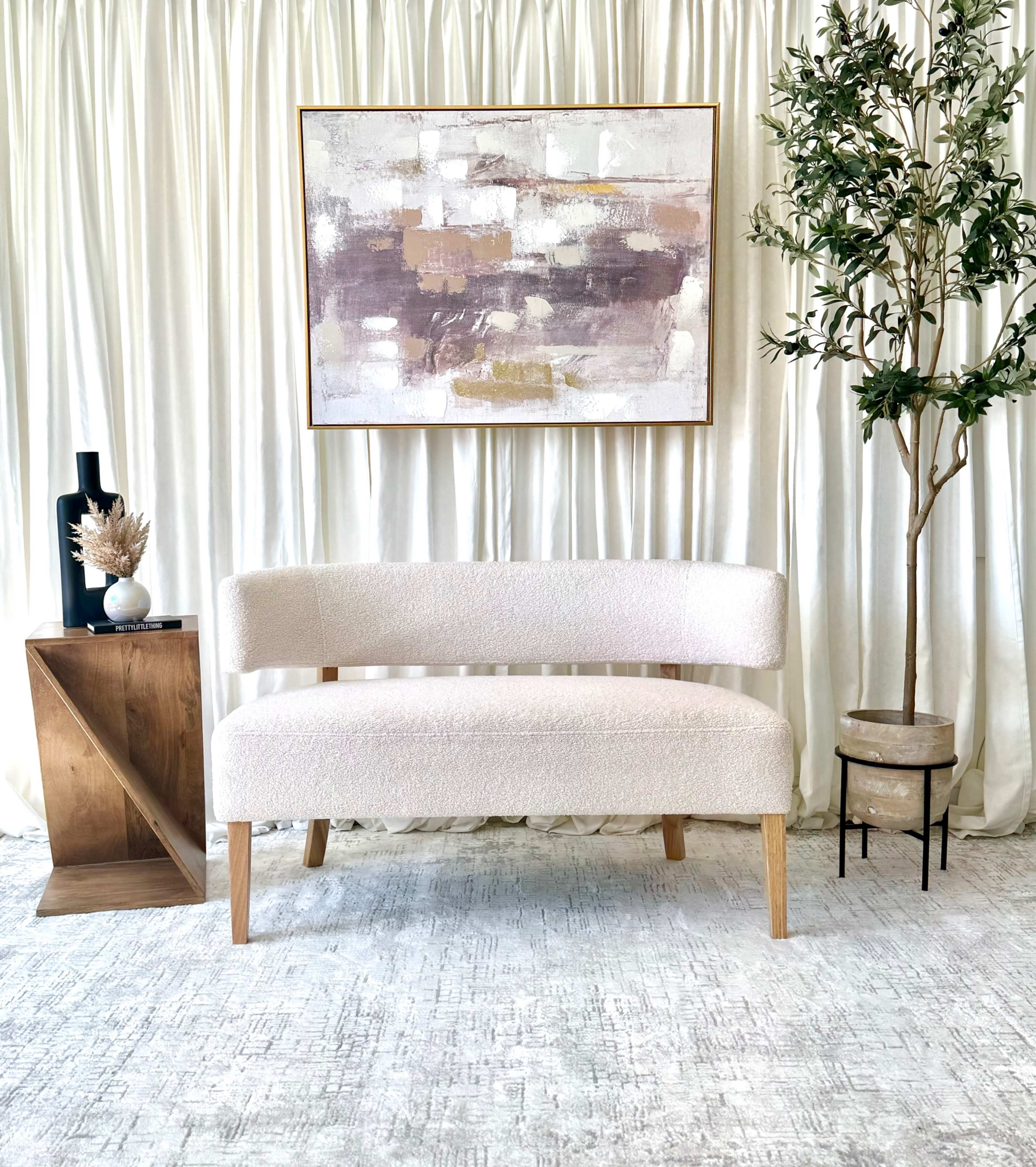 A modern, light-colored bench sits in front of a large abstract painting, accompanied by a wooden side table and a potted plant next to a white curtain backdrop.
