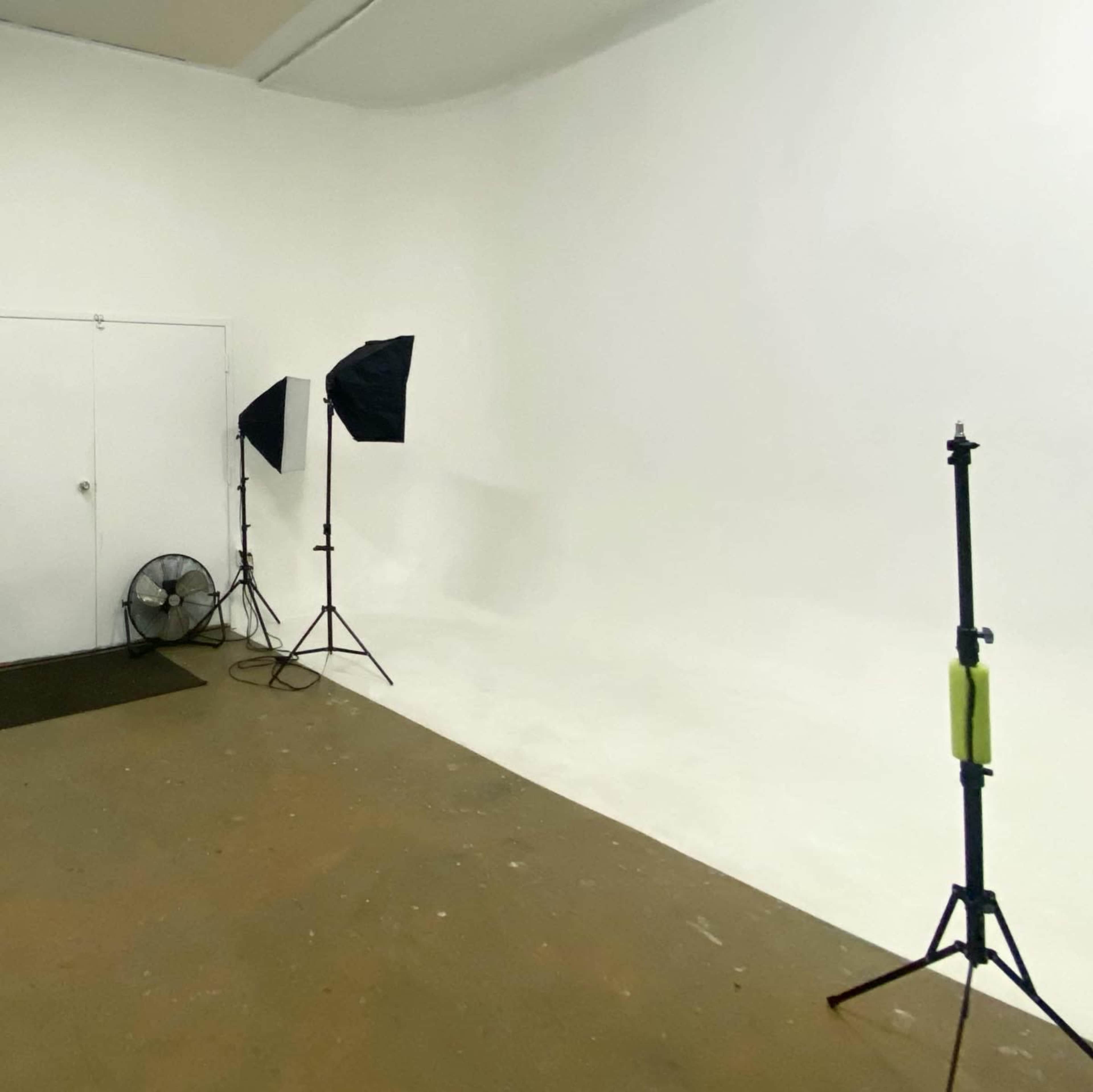 A photography studio features a white backdrop, two softbox lights on stands, a light stand, and a fan in the corner.