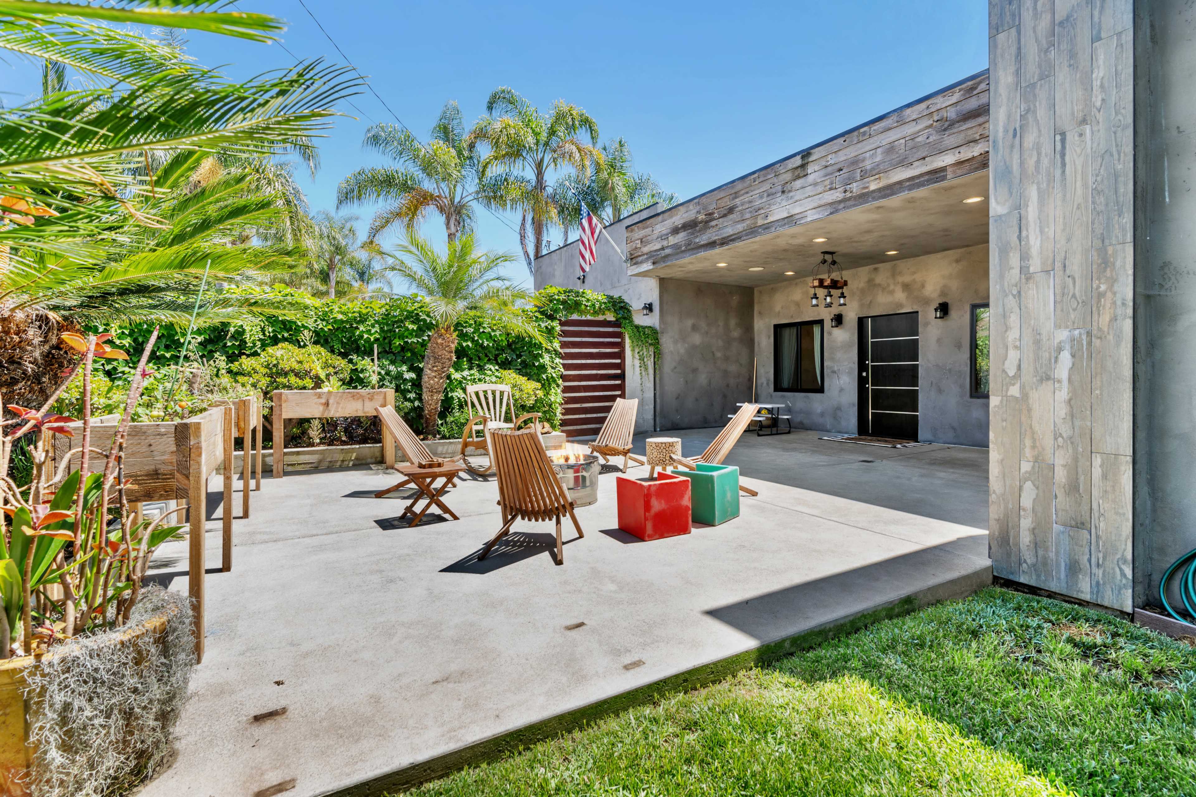 A sunny outdoor patio features wooden chairs, colorful stools, palm trees, and a flag hanging from the roof.