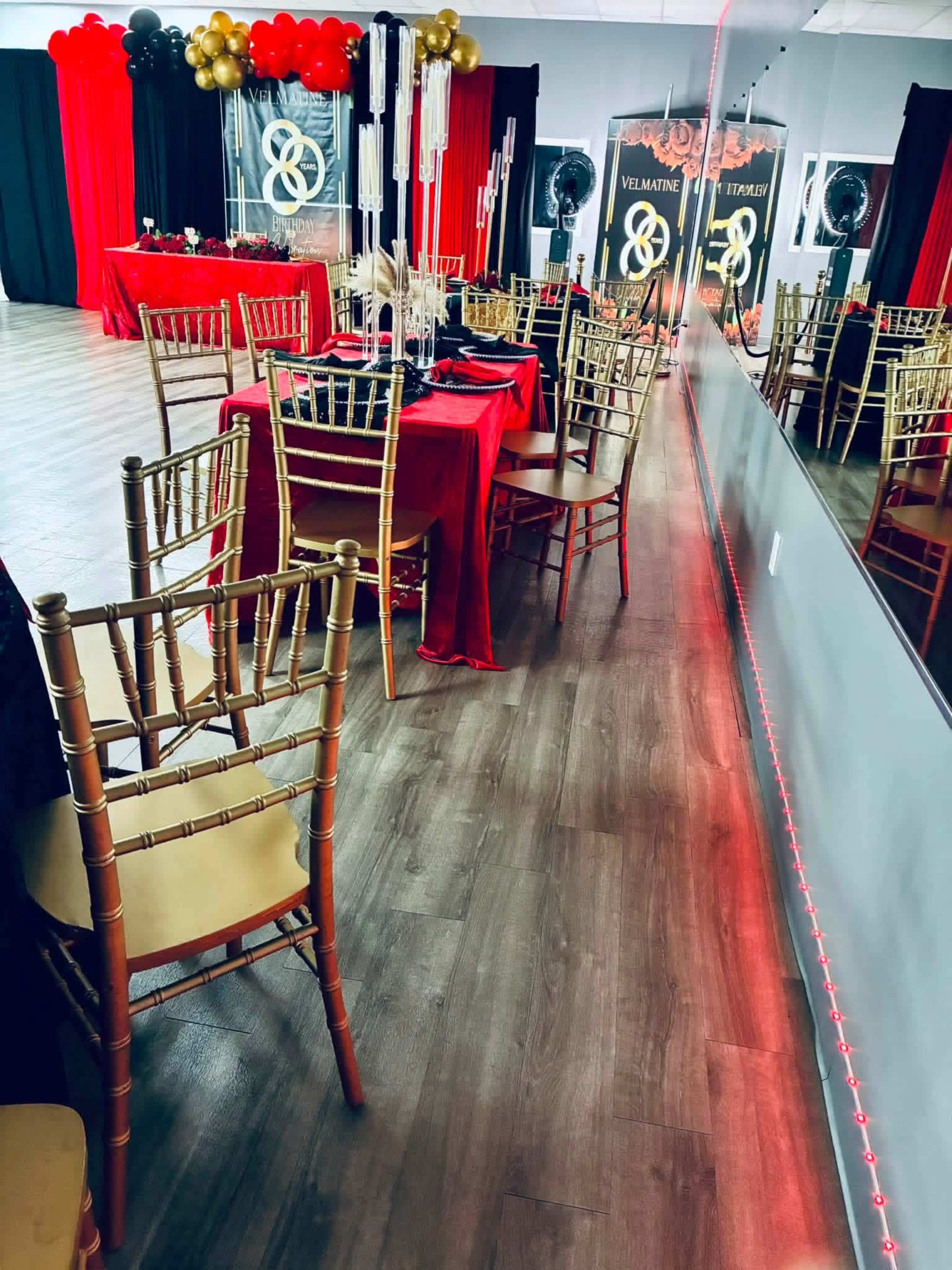 The image shows a decorated event space with gold and black balloons, red tablecloths, and wooden flooring, reflecting the setup in a large mirror.