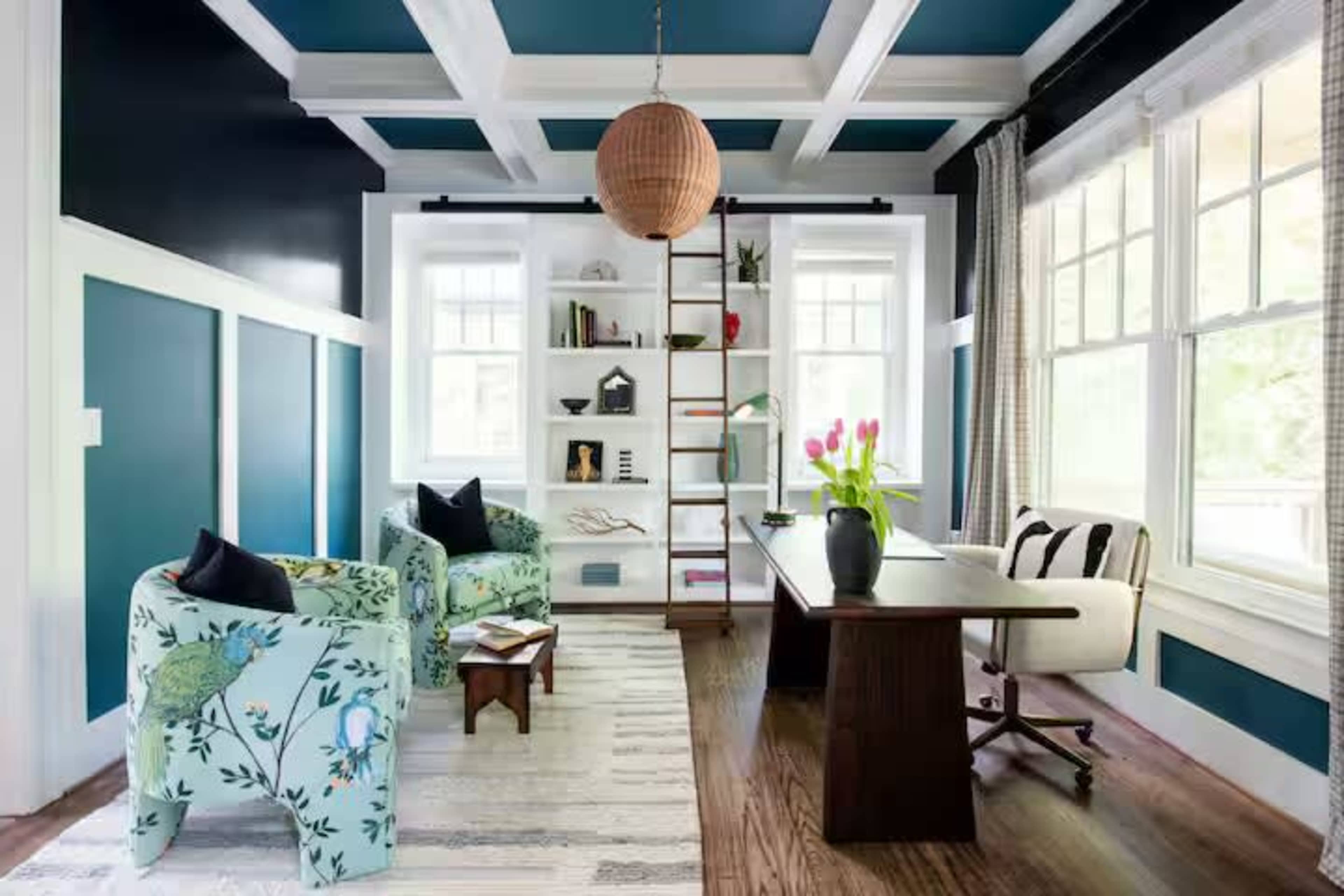 The image shows a modern home office featuring two patterned armchairs, a wooden desk, a ladder bookshelf, and a pendant light, with blue and white walls.