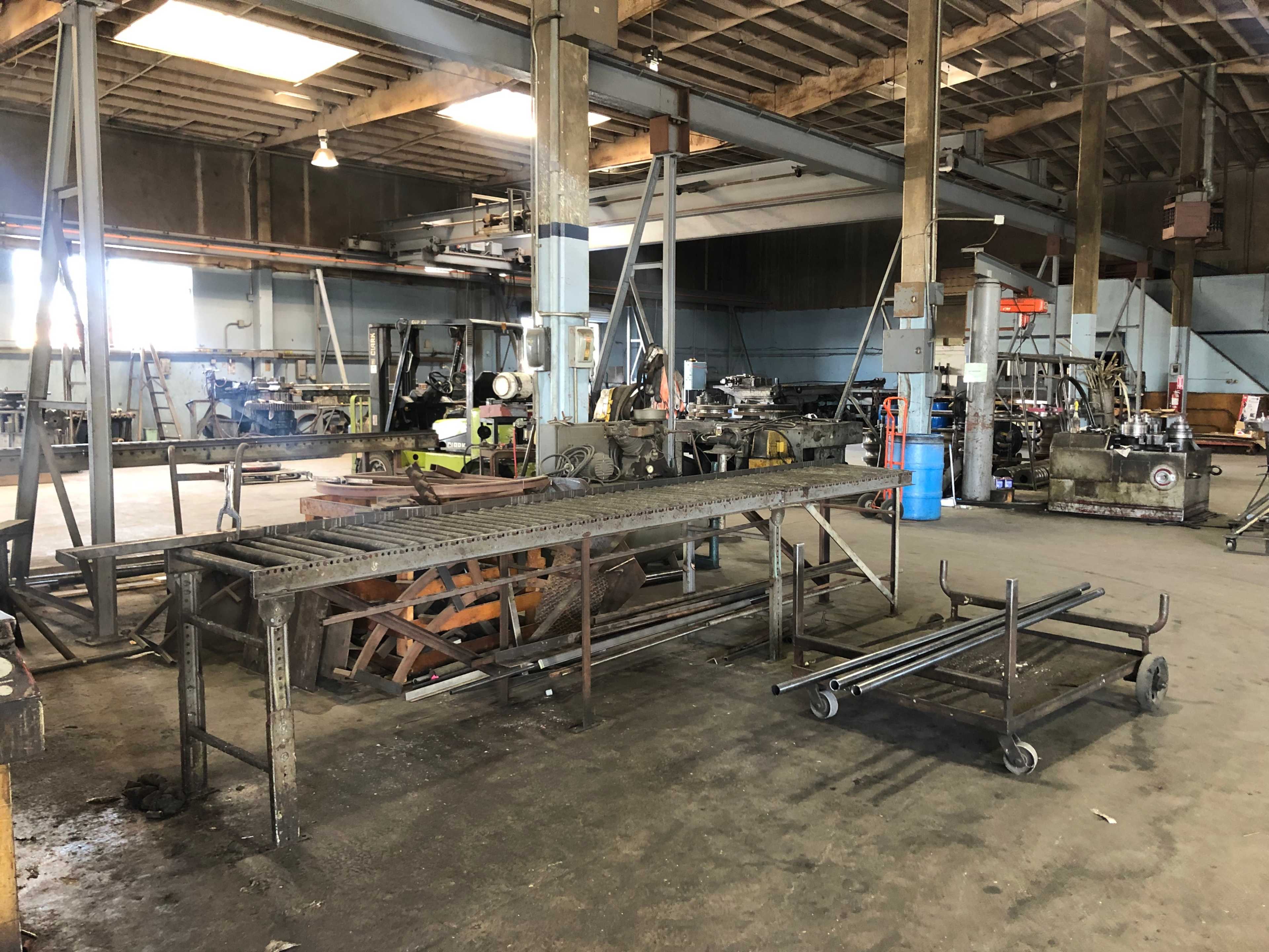 The image shows a spacious industrial workshop with metalworking equipment, storage tables, and tools scattered around a concrete floor.