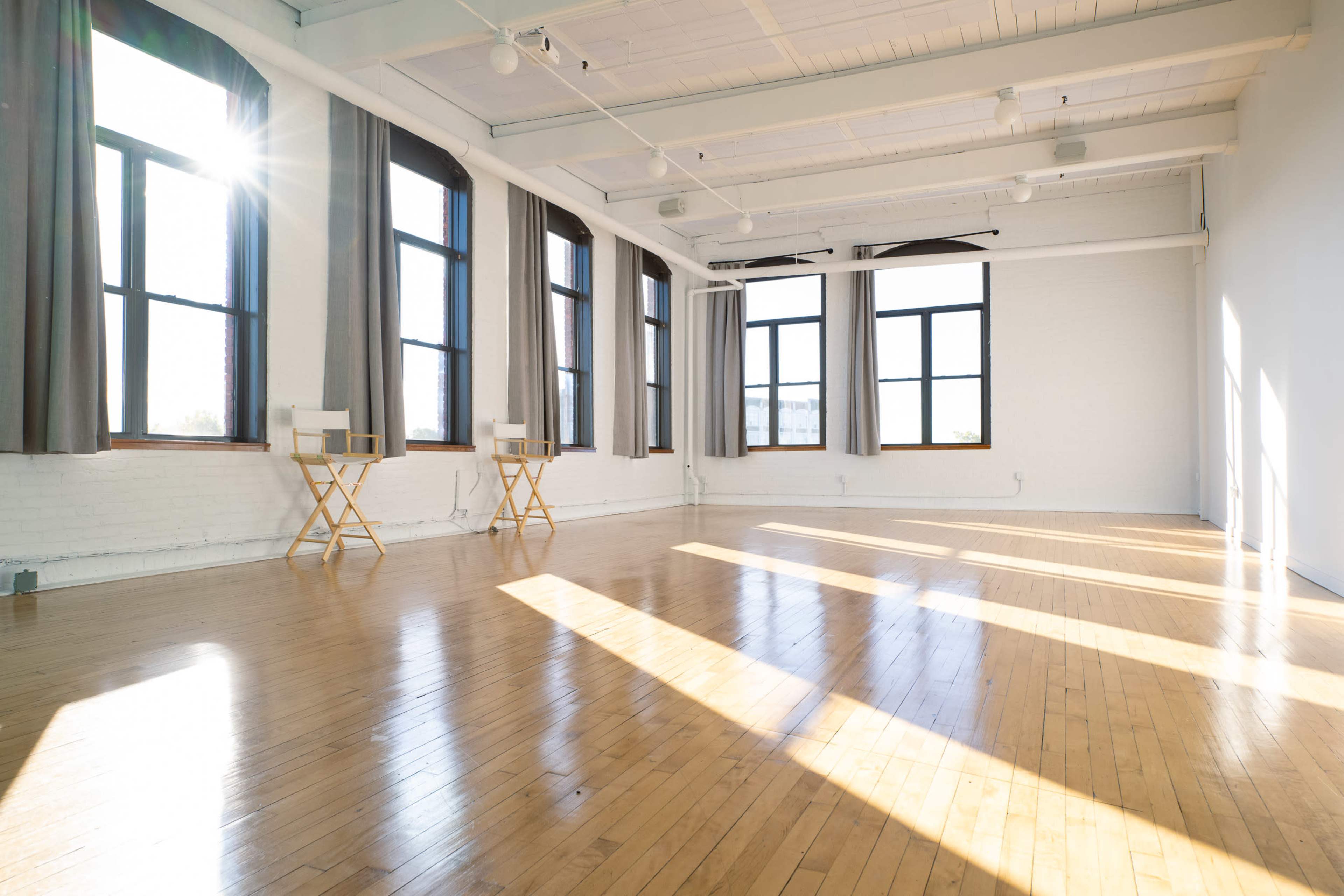 The image shows a bright, spacious room with large windows, wooden floors, and minimal furnishings.