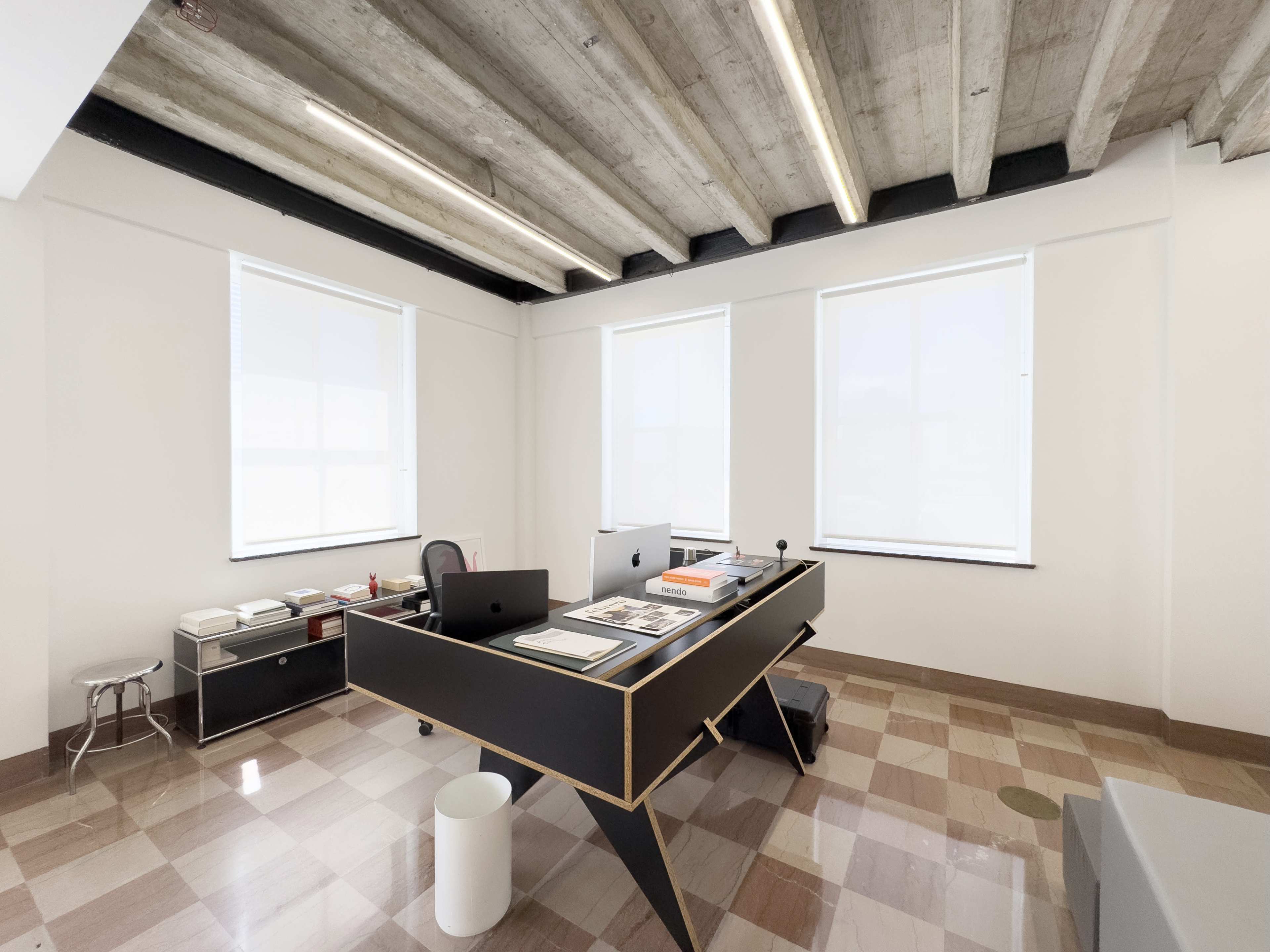 The image depicts a modern office space with a black desk, two computers, and large windows allowing natural light to enter.
