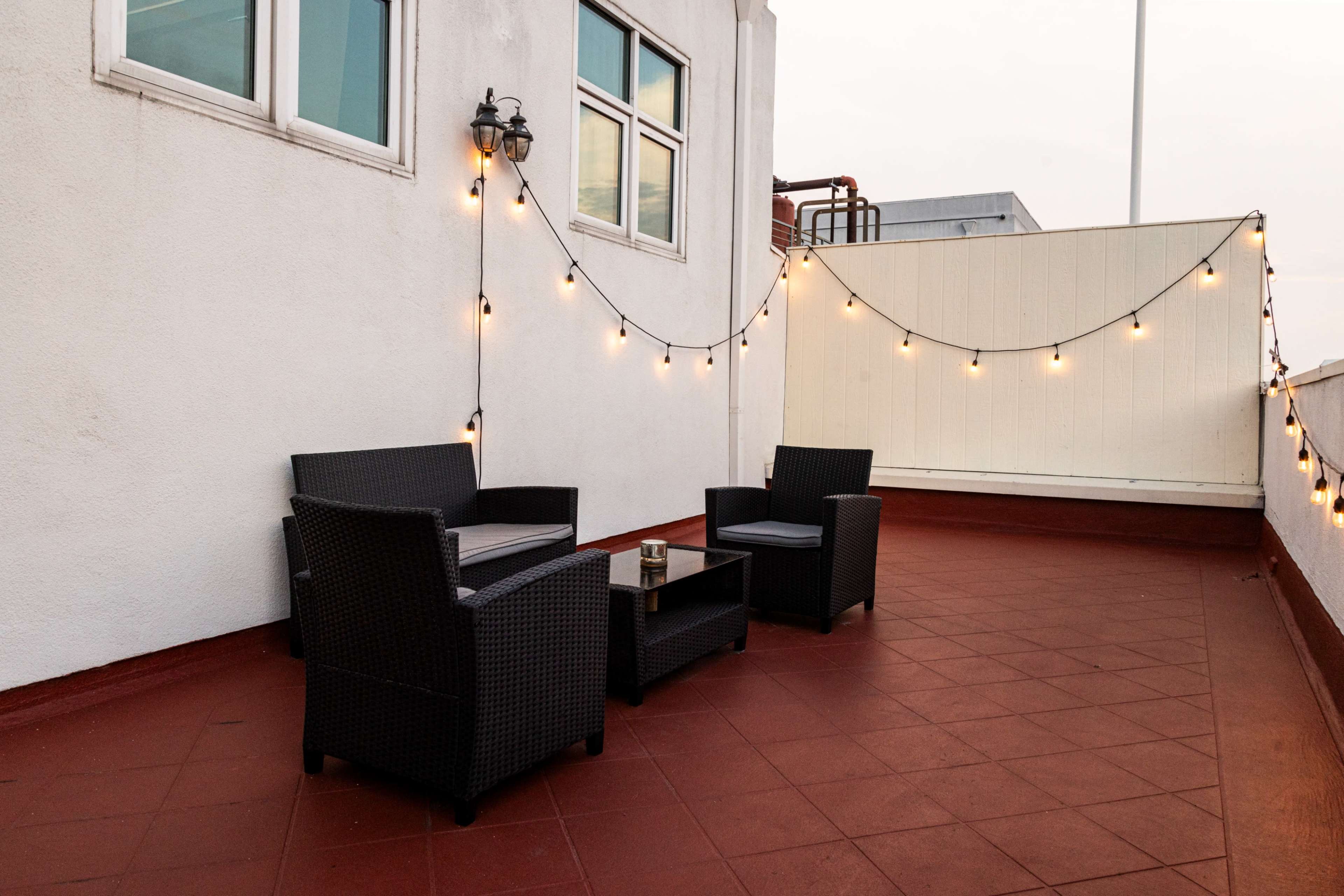 The image shows a rooftop terrace with two black wicker chairs, a small table in between, and string lights hanging along the white wall.