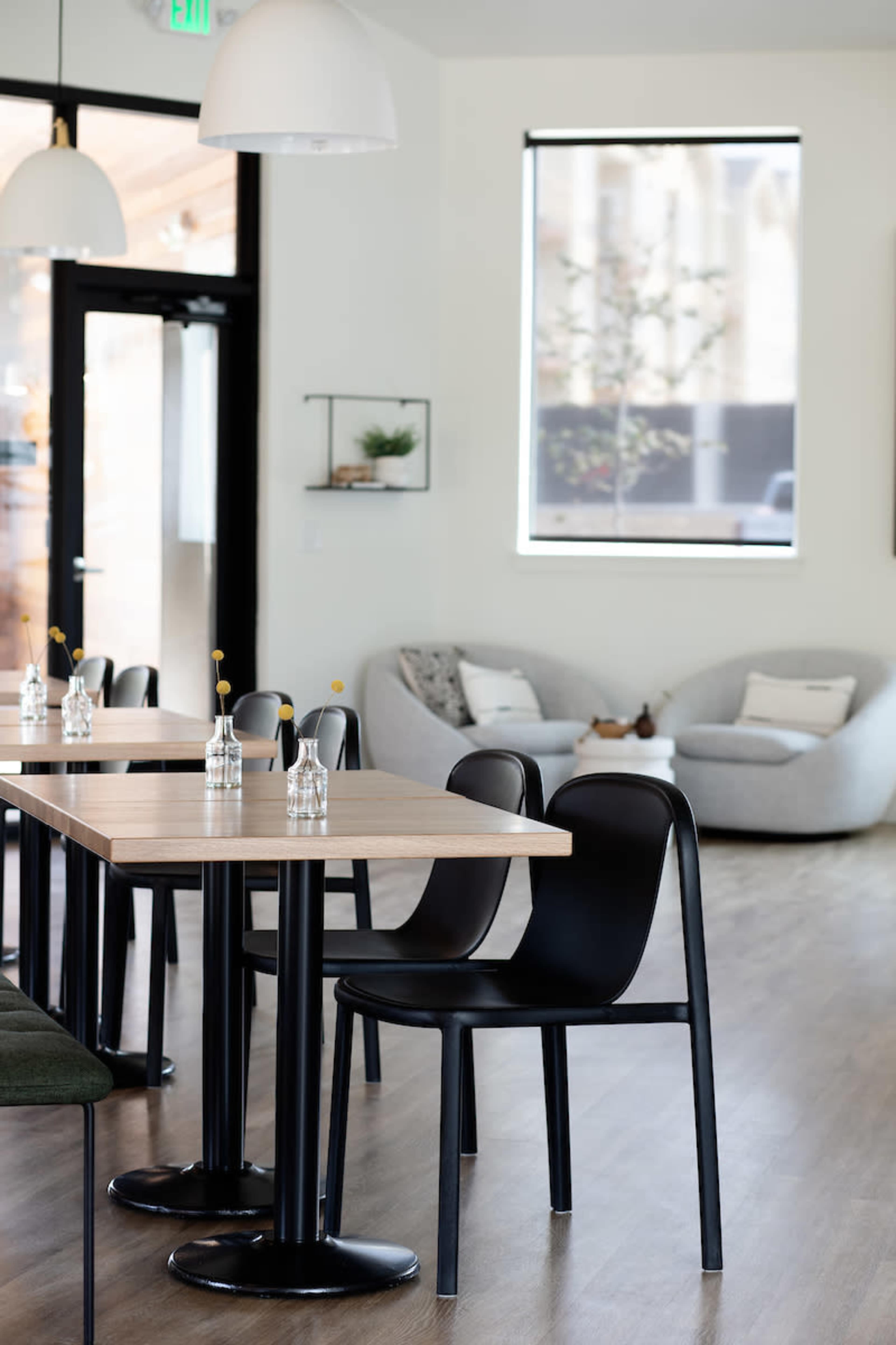 The image shows a modern, minimalist café interior with wooden tables, black chairs, and a cozy seating area in the background.